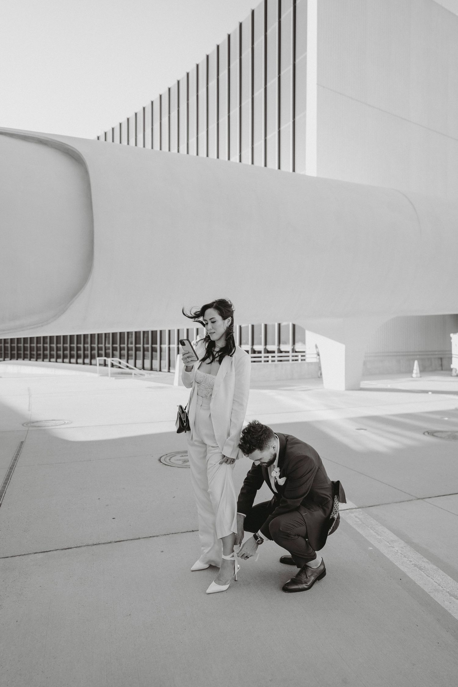 Groom kneels to fix bride's shoe outside the iconic curved white exterior of the TWA Hotel at JFK Airport.