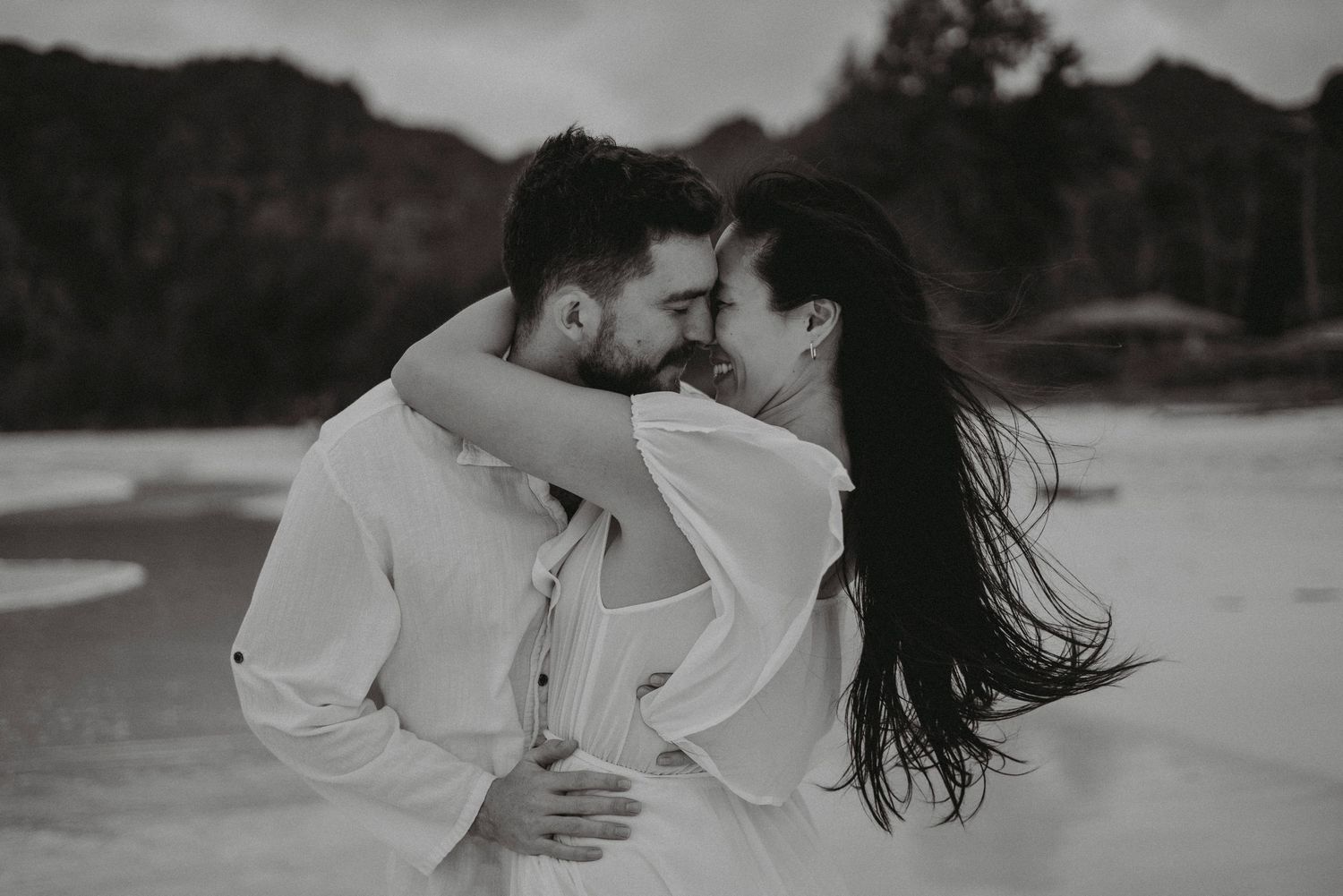 Black and white photo of a couple nearly kissing on a windy beach, wearing white outfits.