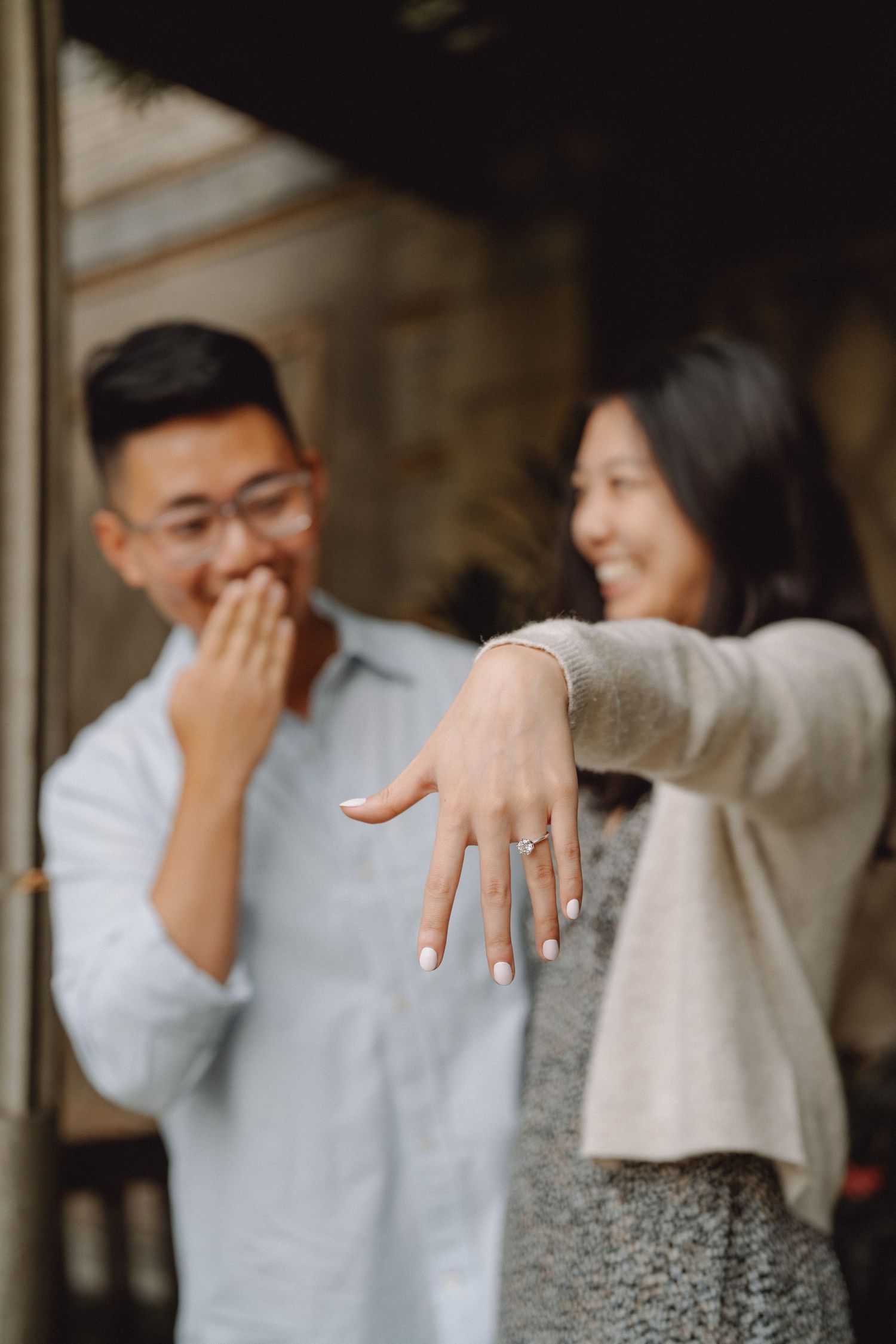 Smiling woman holds out her hand to show a new engagement ring while her happy fiancé covers his mouth in joyful surprise.