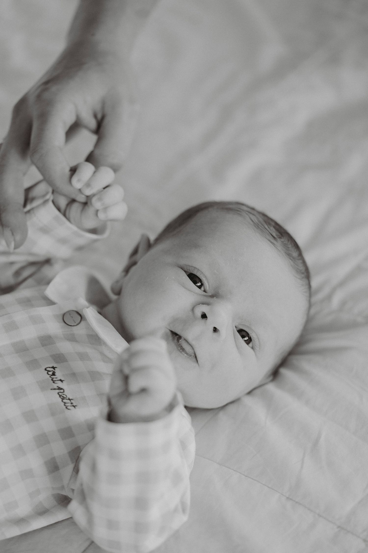 Black and white photo of alert newborn baby holding parent's finger on bed.
