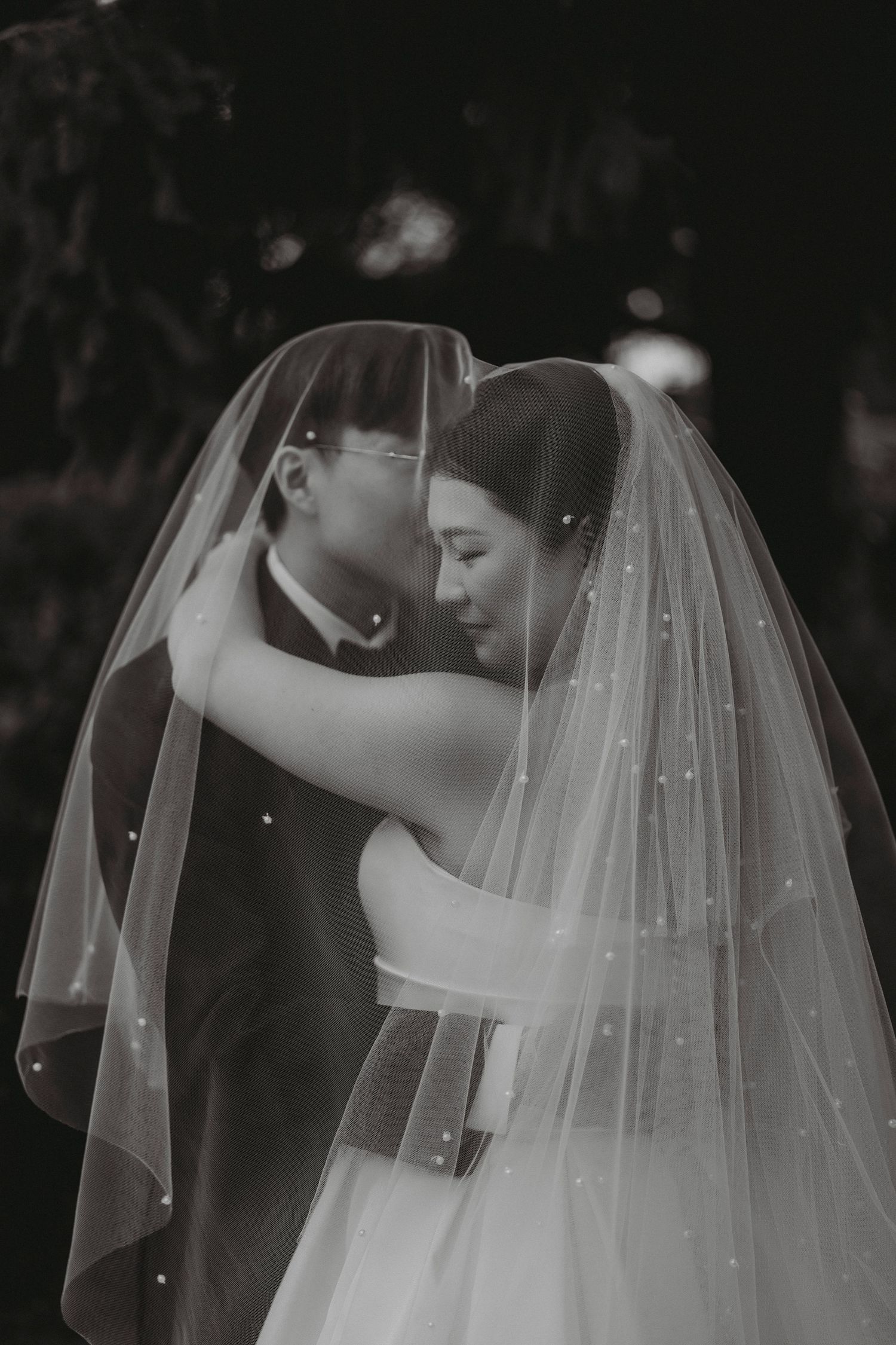 Bride and groom tenderly embrace under flowing pearl veil in romantic black and white portrait.