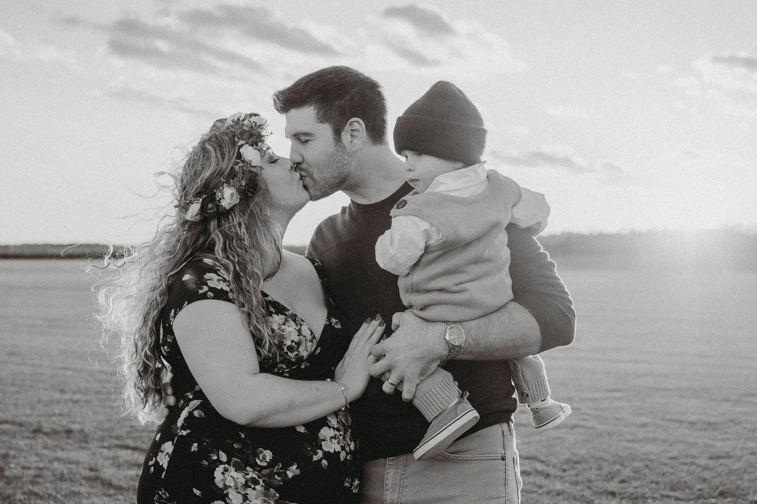 Black and white photo of a couple sharing a kiss while holding their baby in an open field at sunset.