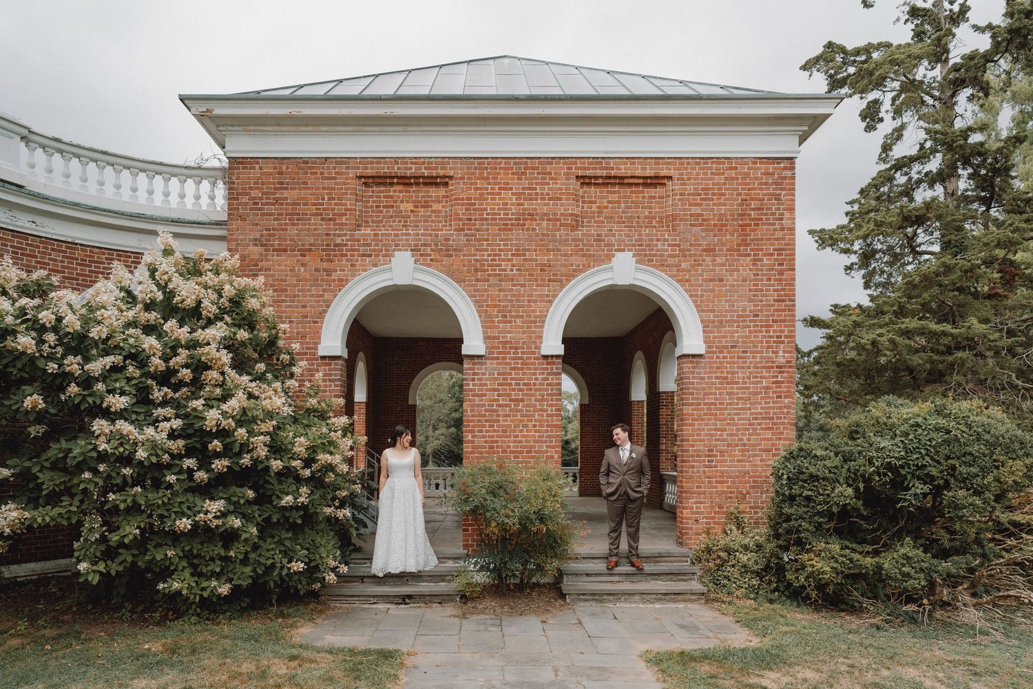 Bride and groom standing apart at arched doorways of historic brick building on wedding day.