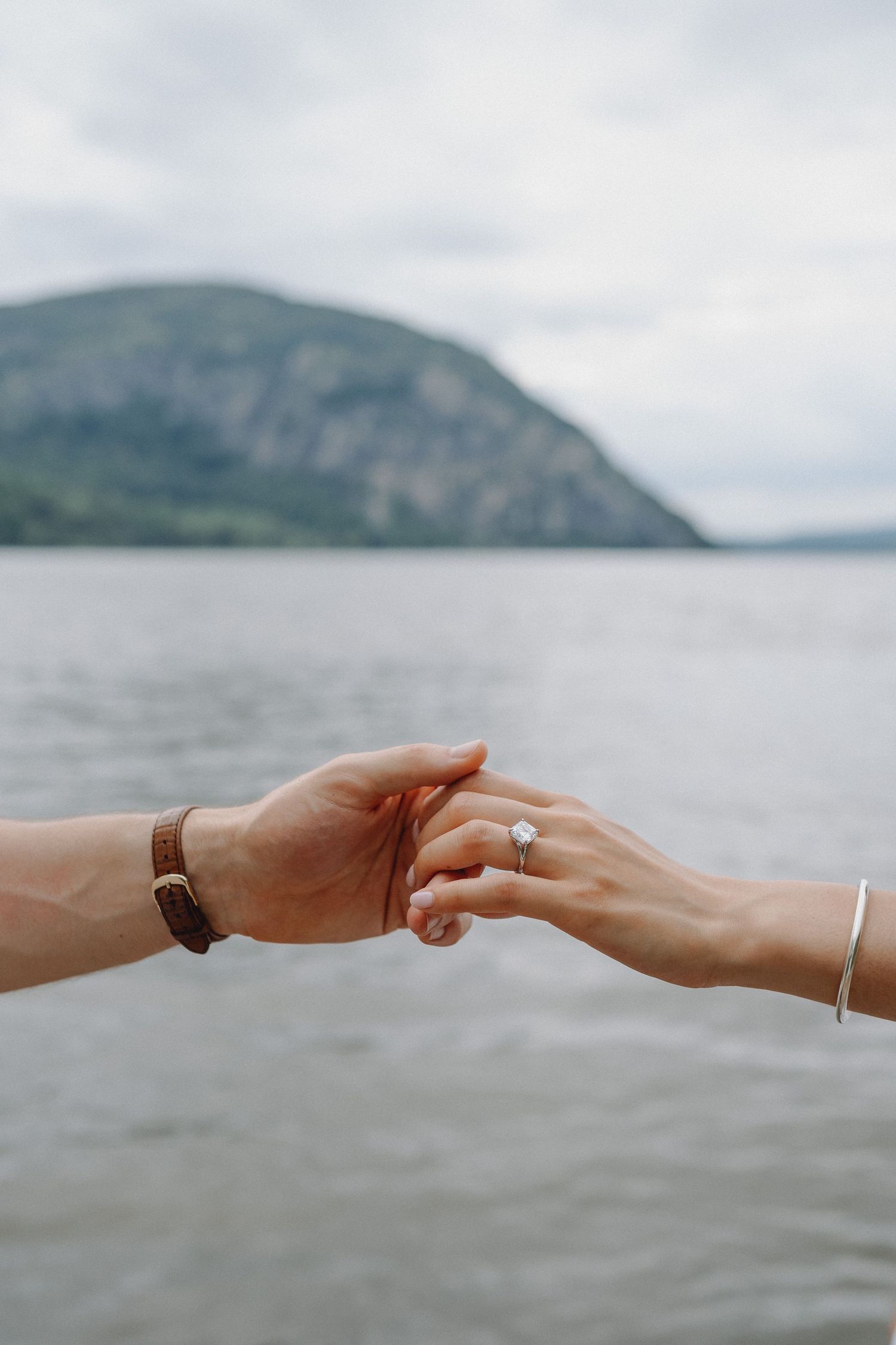 Two hands gently touching over a calm lake, one wearing a leather watch, the other a diamond engagement ring, with a mountain behind.