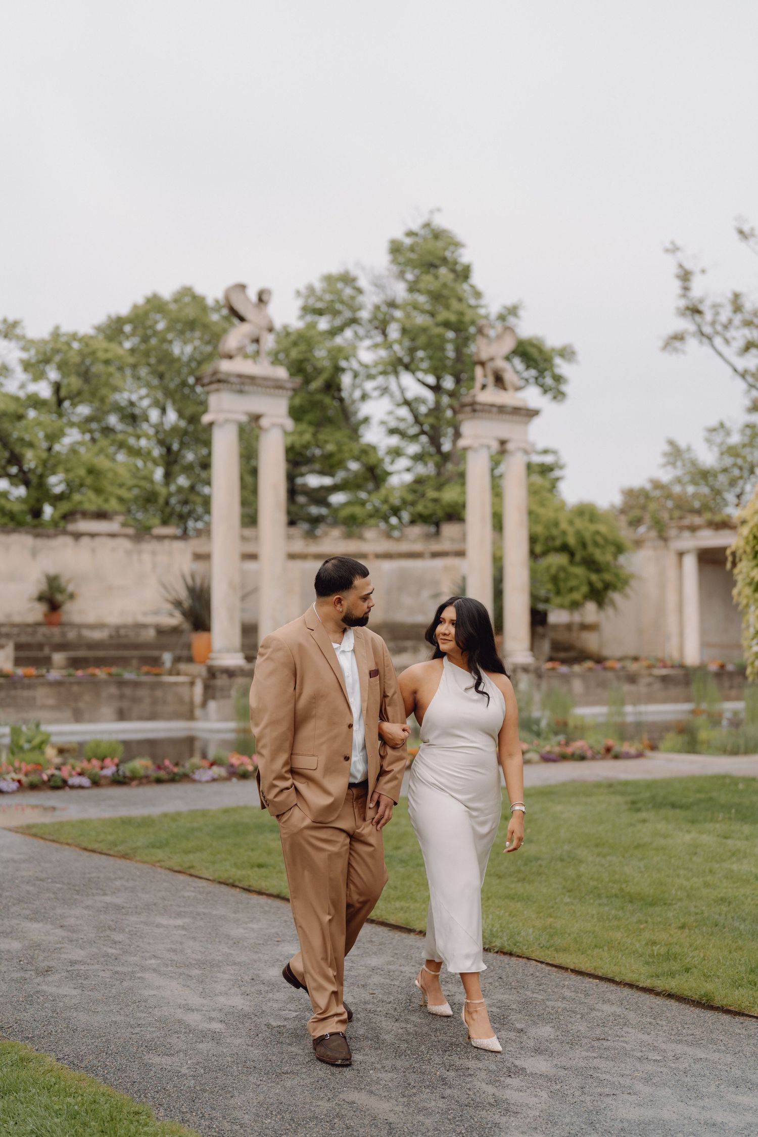 Couple walking hand in hand through a formal garden with classical stone columns and lush greenery.