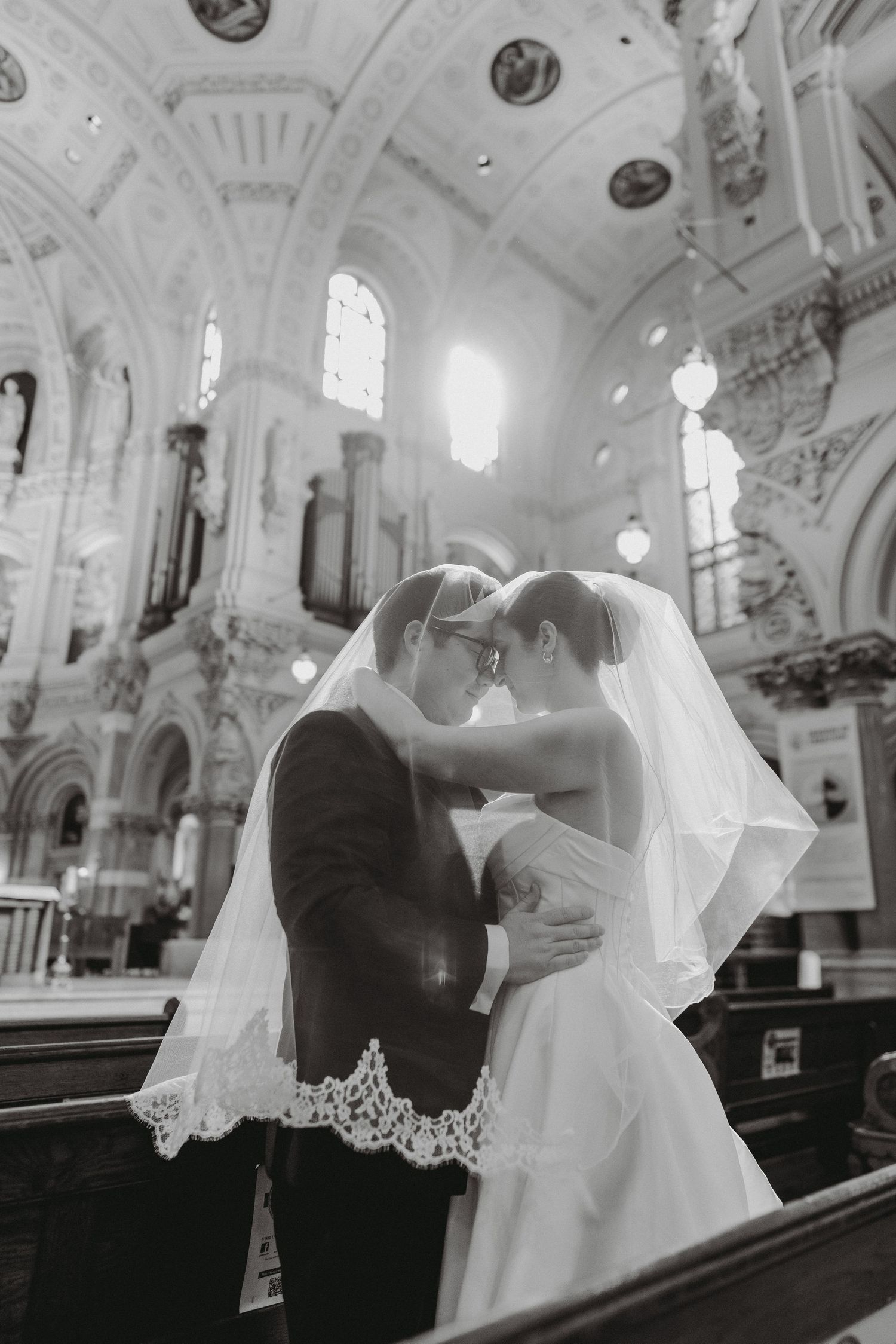 Black and white romantic photo of bride and groom touching foreheads under veil in church.