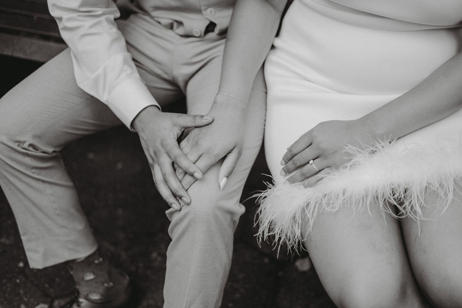 Black and white close-up of a couple holding hands on a bench, woman wearing engagement ring and feathered skirt.