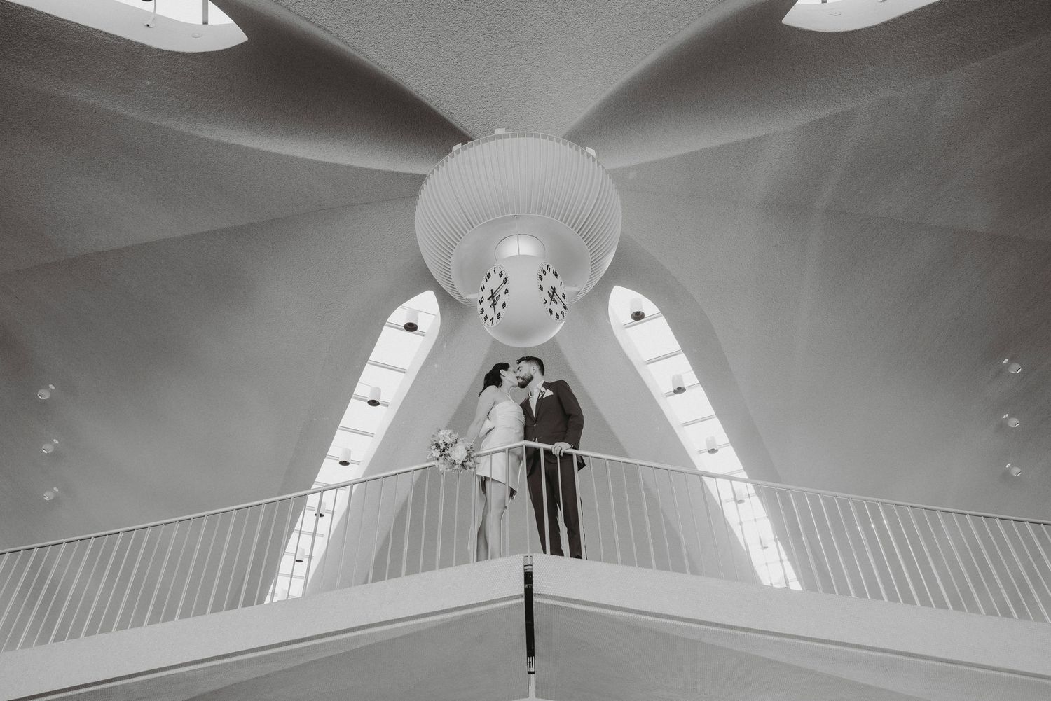 Bride and groom share a kiss on the sweeping staircase beneath the dramatic vaulted ceiling of the TWA Hotel.