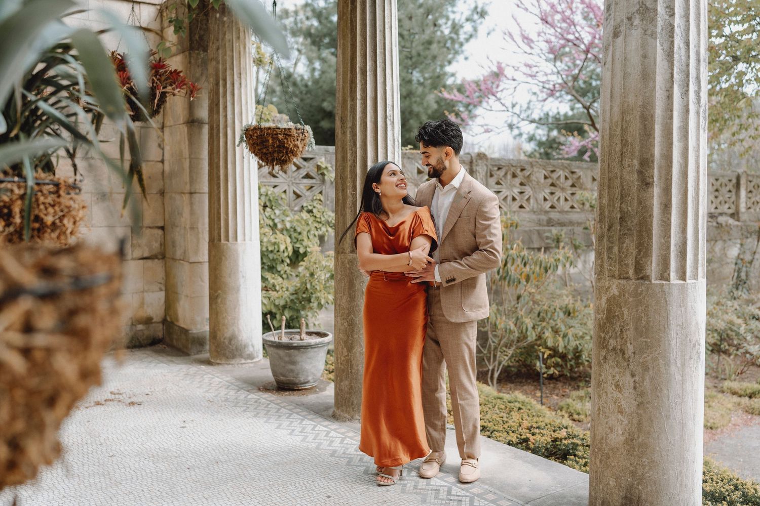Couple in rust orange dress and beige suit embracing among stone columns in a garden setting.