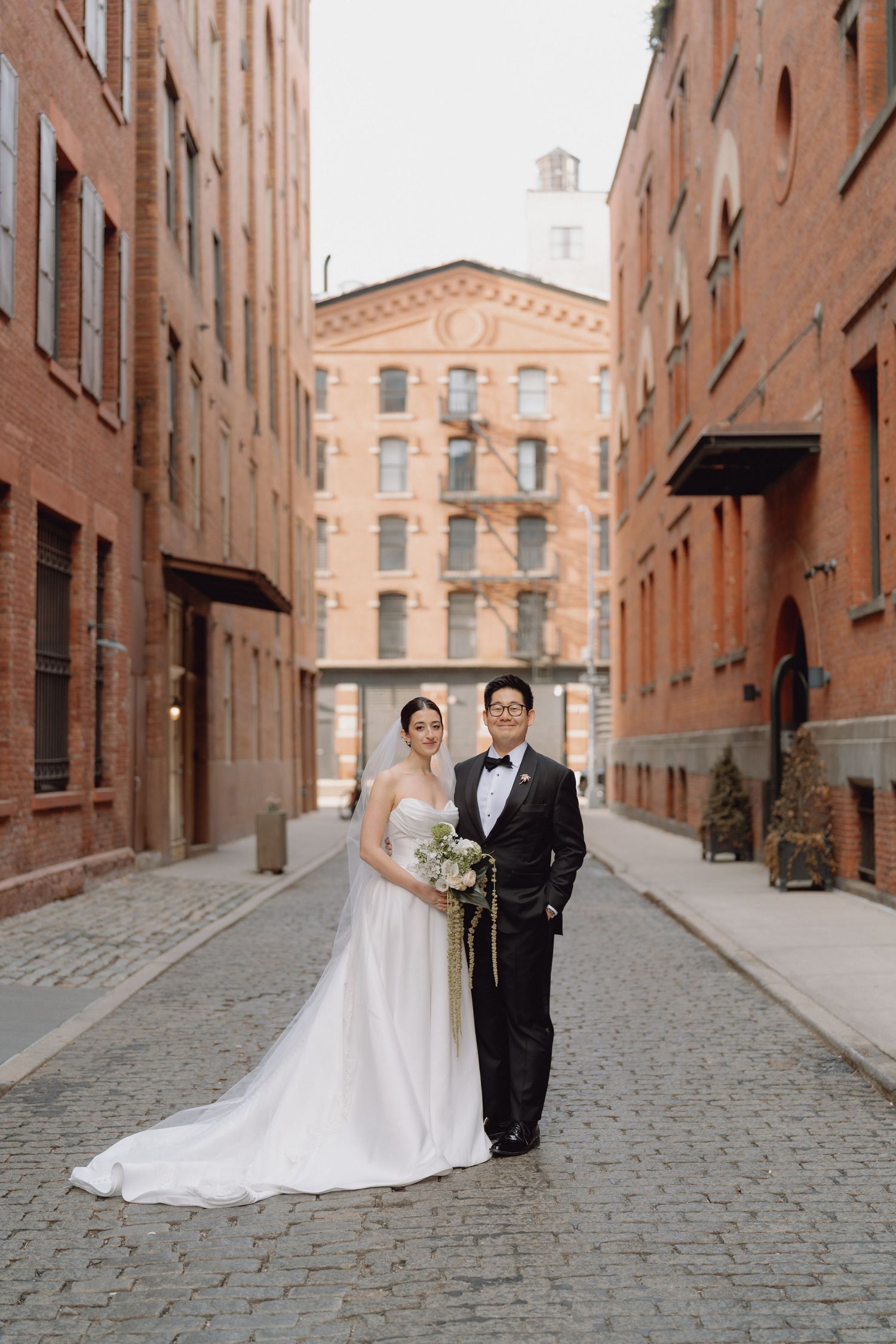 Bride and groom pose on cobblestone alley between historic red brick buildings on their wedding day.