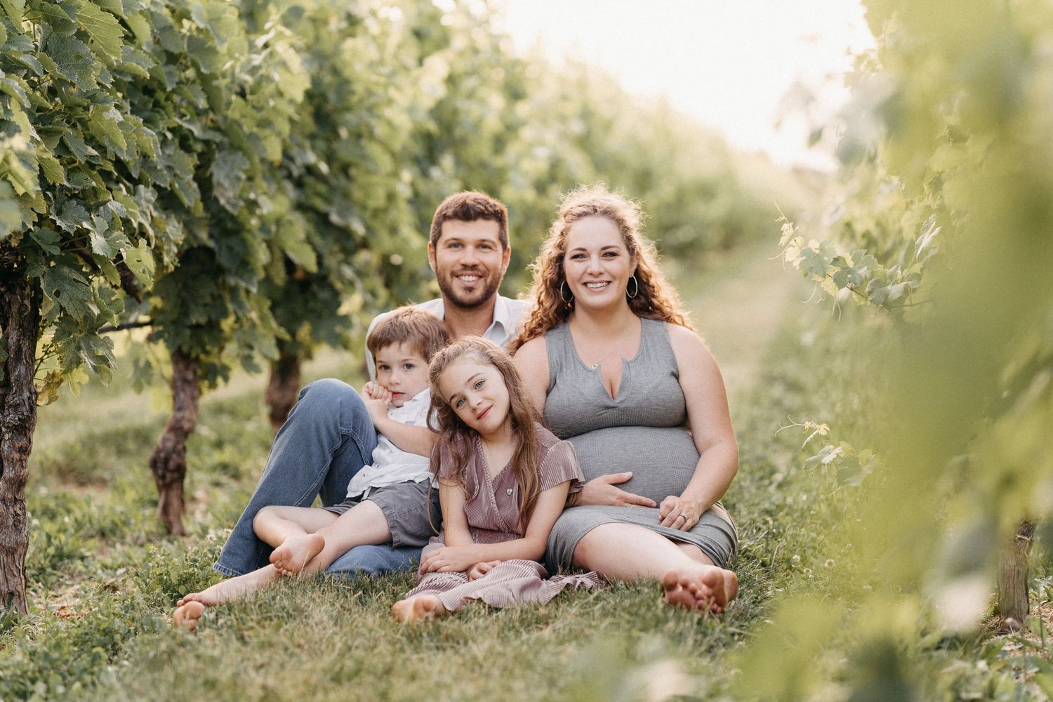 A smiling family of four sits together in a lush green vineyard during golden hour.