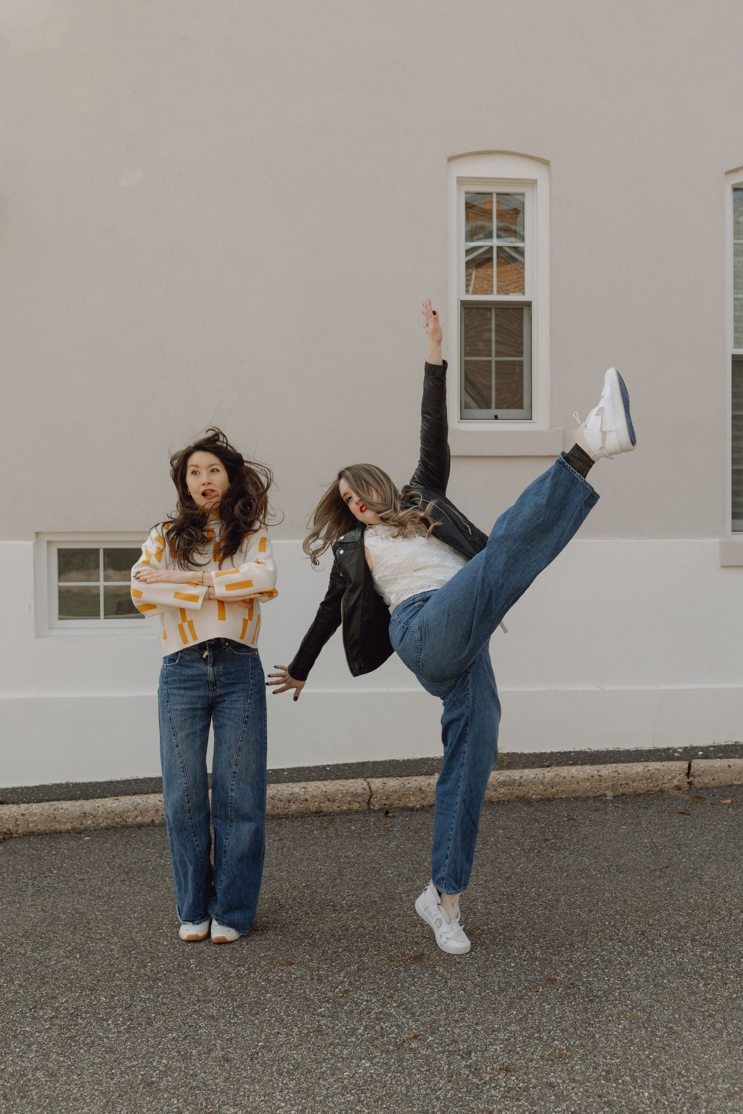Two women posing outside building, one doing a high kick in jeans and white sneakers while other watches.
