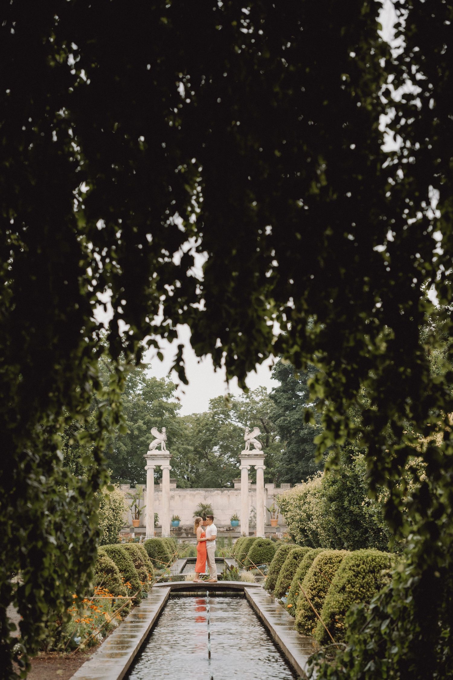 Couple in coral and white standing by a long reflecting pool in a formal garden with trimmed hedges, stone pillars and statues.
