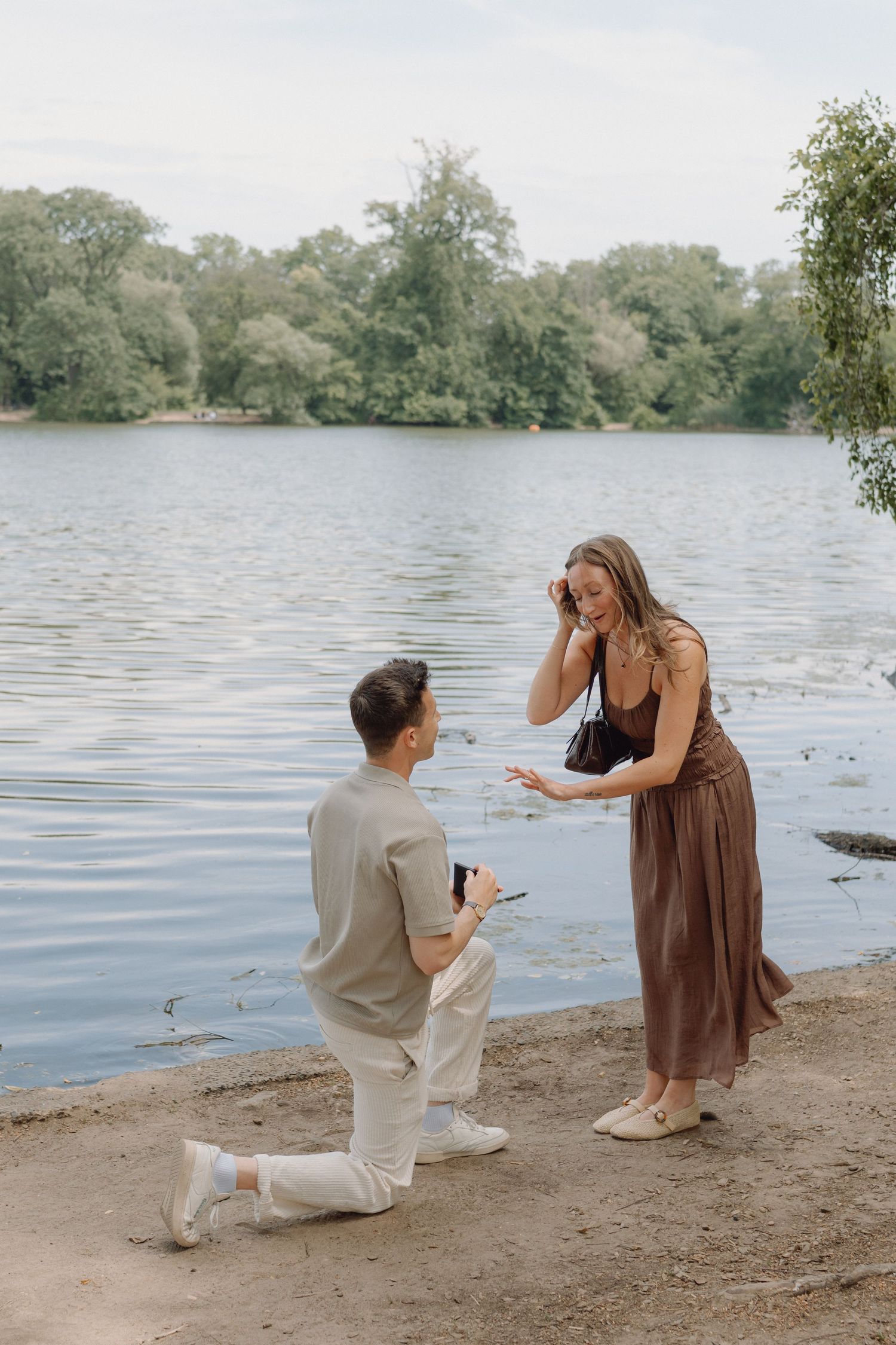 Man kneeling on one knee proposes to a smiling woman in a brown dress beside a calm lake with trees in the background.