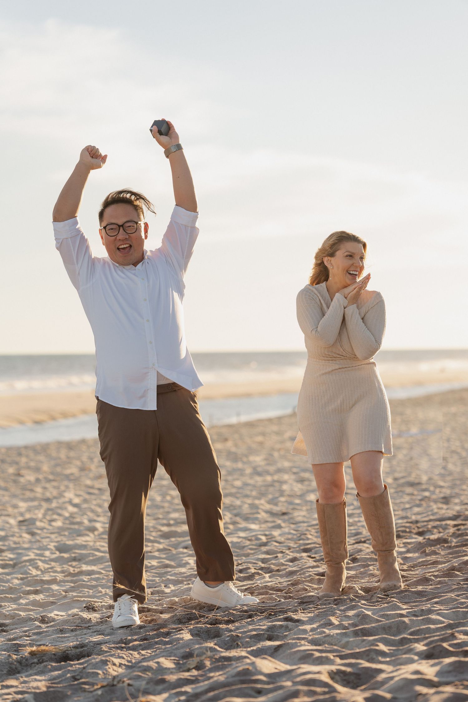 Joyful man with raised arms cheers beside a smiling woman clapping her hands on a sunlit sandy beach at sunset.