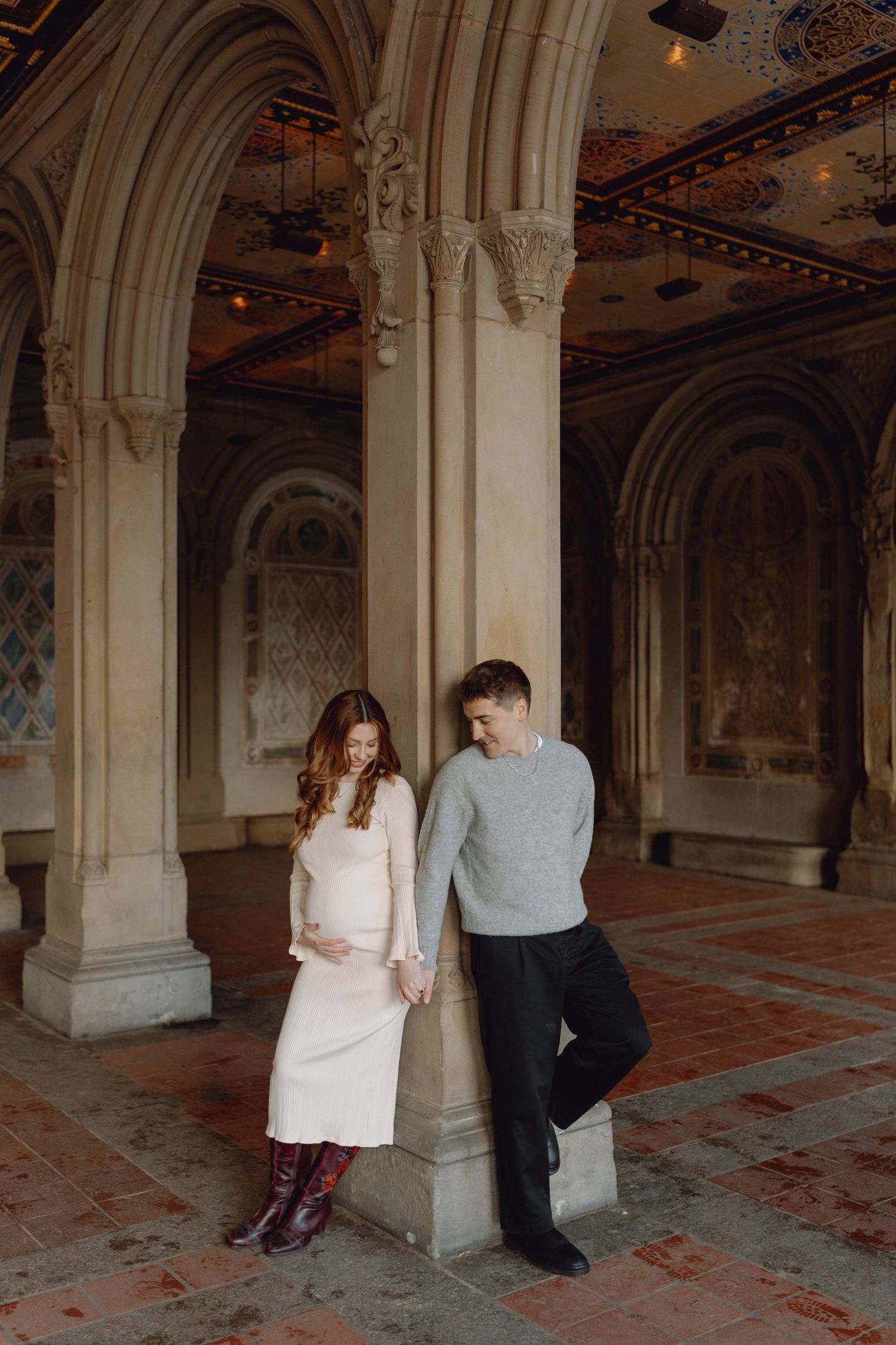 Pregnant woman in white dress holding belly with partner under ornate stone archway.