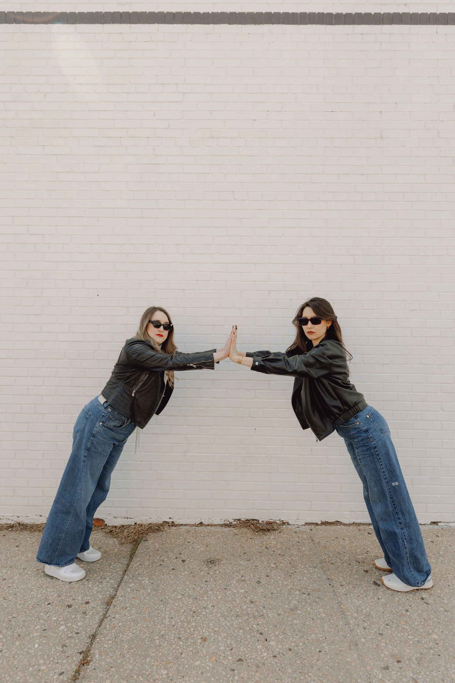 Two women in black leather jackets and jeans facing each other with hands touching against a white brick wall.