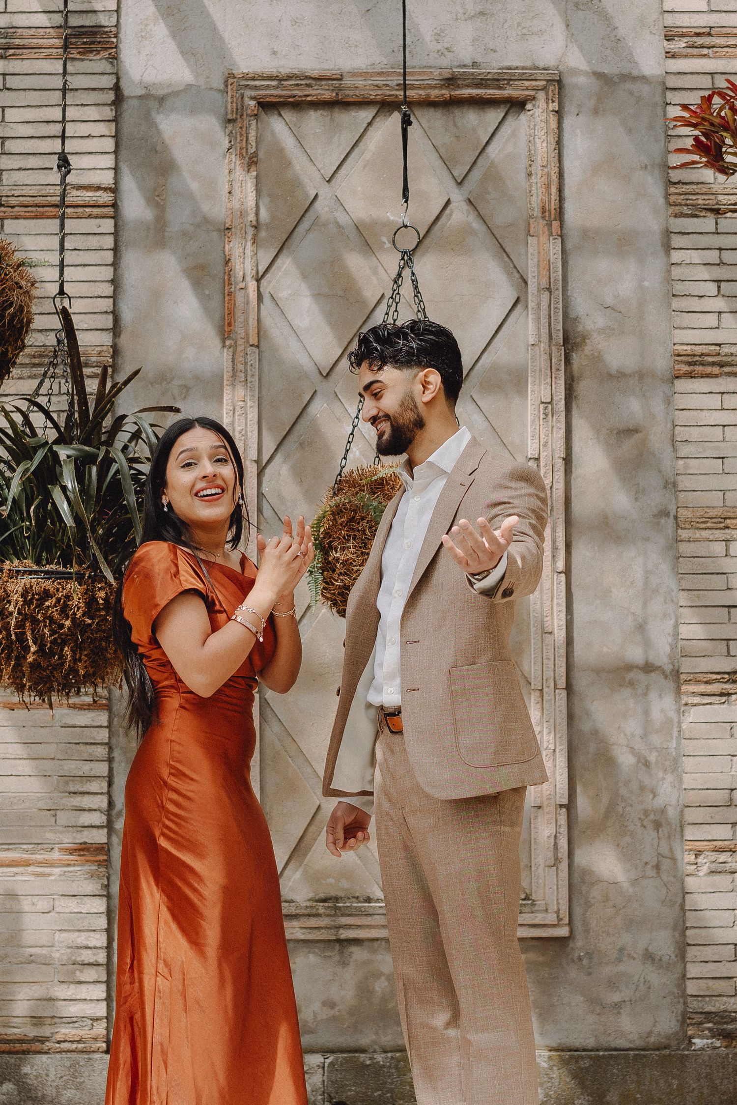 Smiling couple in rust dress and beige suit celebrate joyfully beside hanging plants and lattice wall.