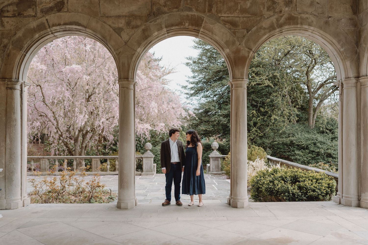 Couple standing under elegant stone arches with blooming cherry blossom trees in background.