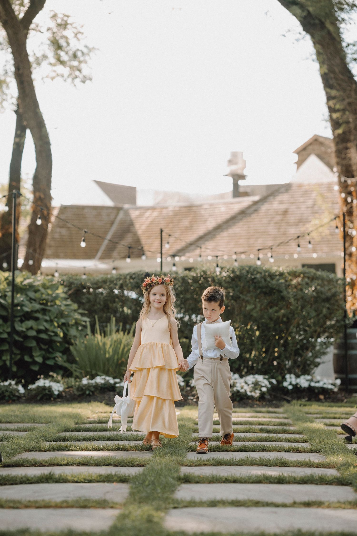 Flower girl in yellow ruffled dress and ring bearer in tan suit walk down stone aisle at outdoor wedding.