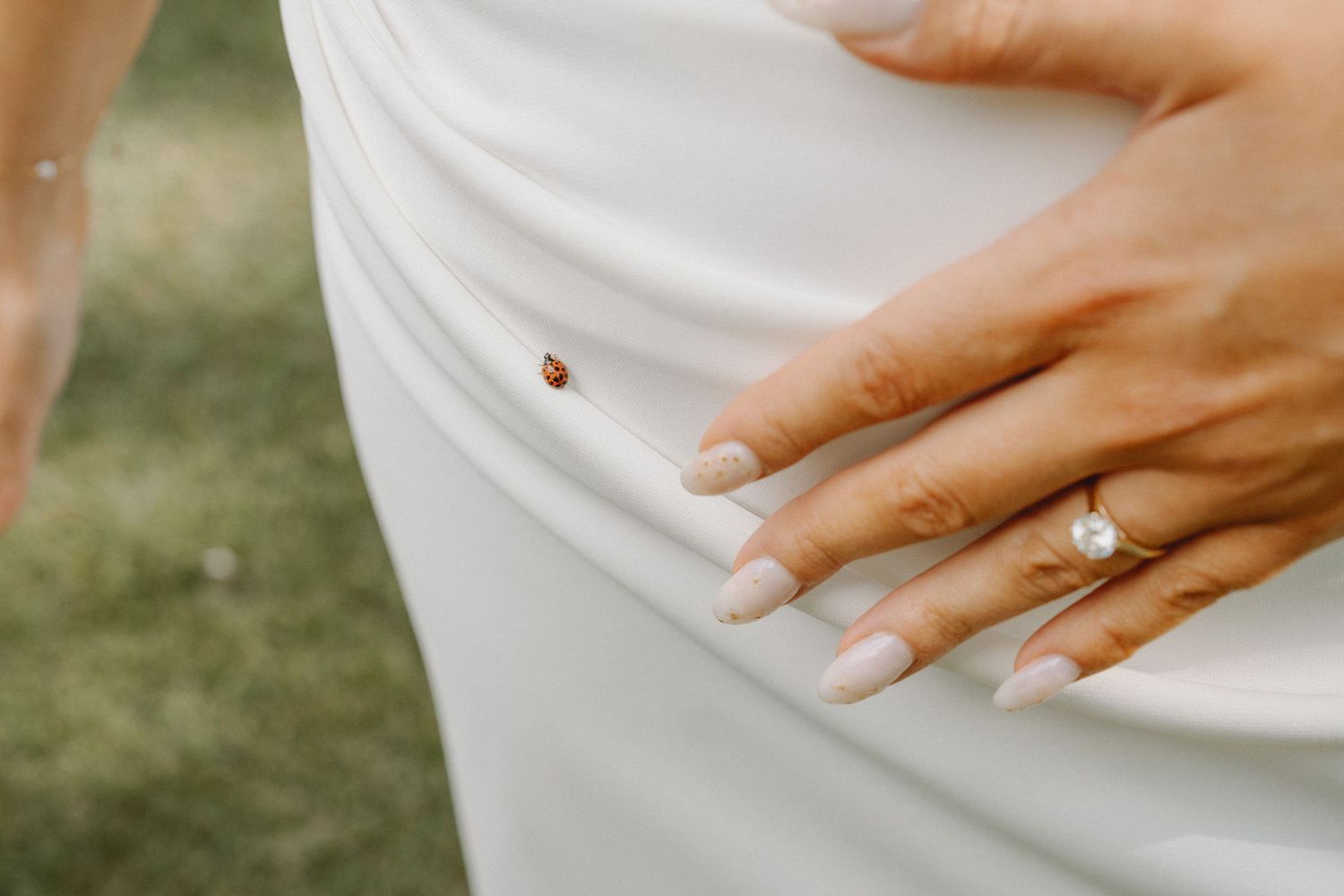 Close-up of bride's diamond engagement ring and manicured nails with ladybug on white wedding dress.
