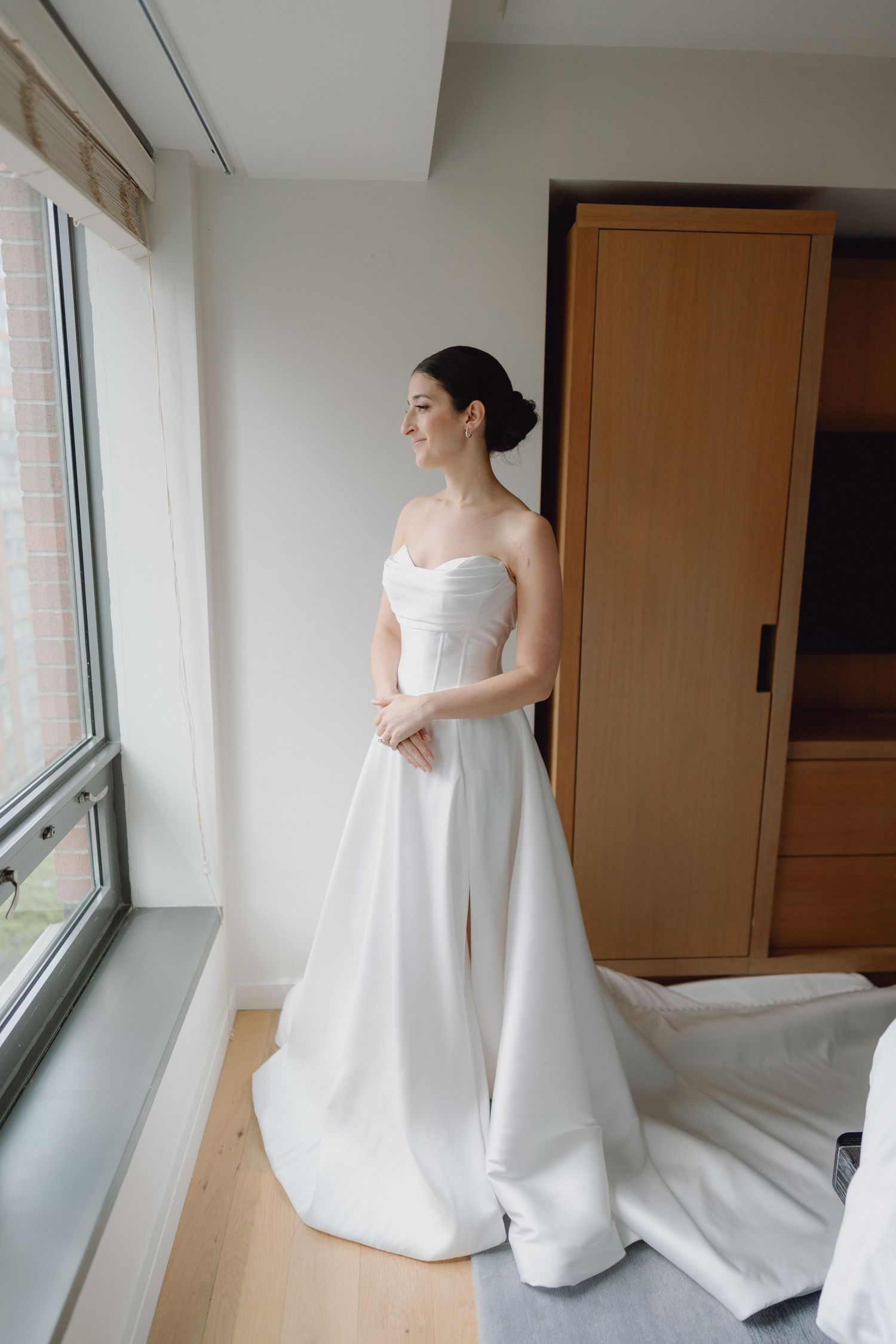 Elegant bride in strapless white satin wedding gown stands by a window in a bright hotel room.