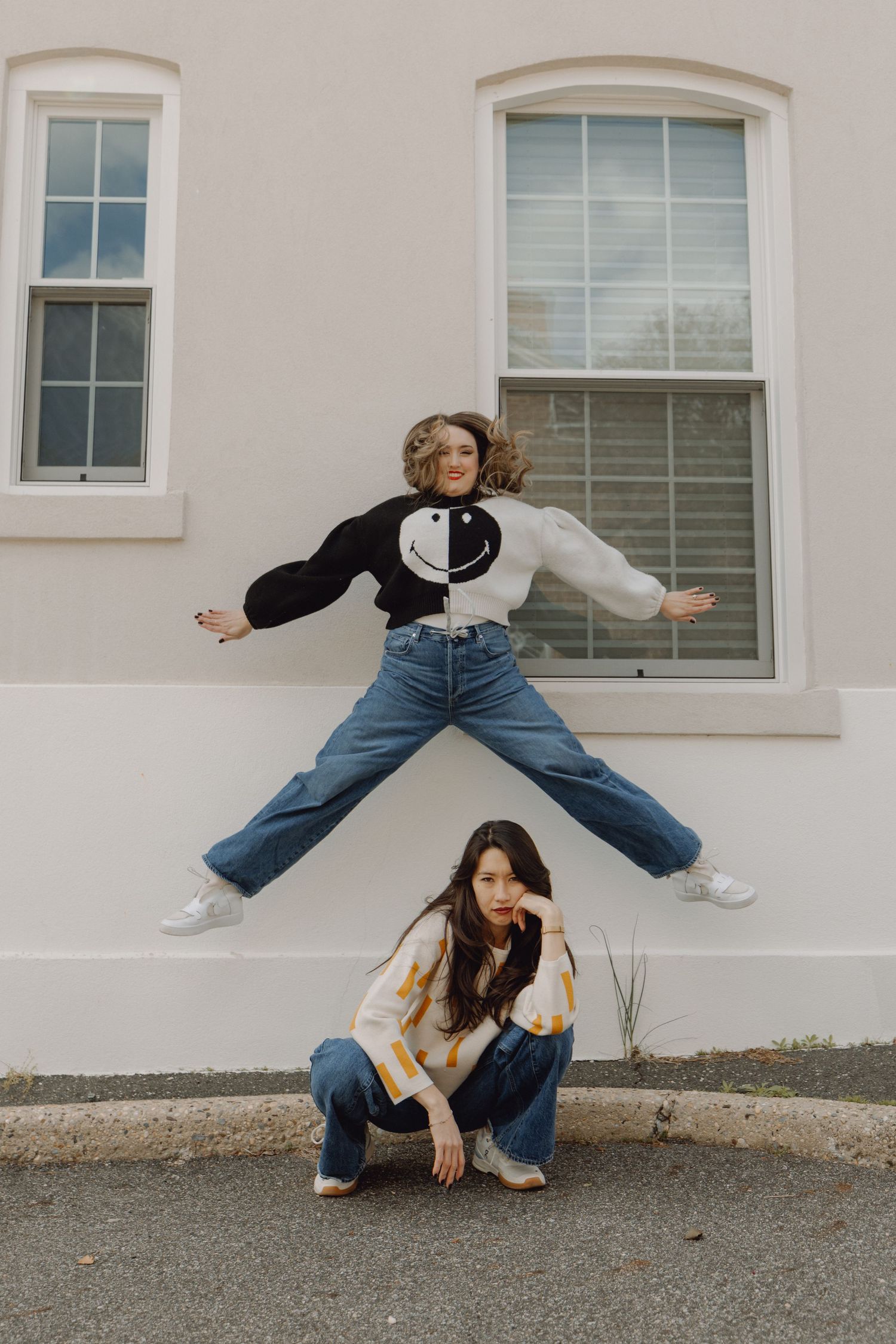 Two women posing outside, one jumping high in a smiley face sweater while the other crouches below.