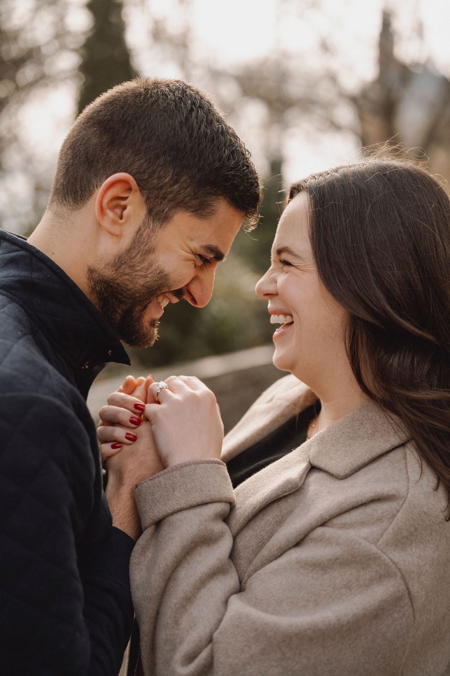 Smiling couple holding hands and laughing face to face outdoors, with the woman wearing a ring in a romantic engagement-style photo.