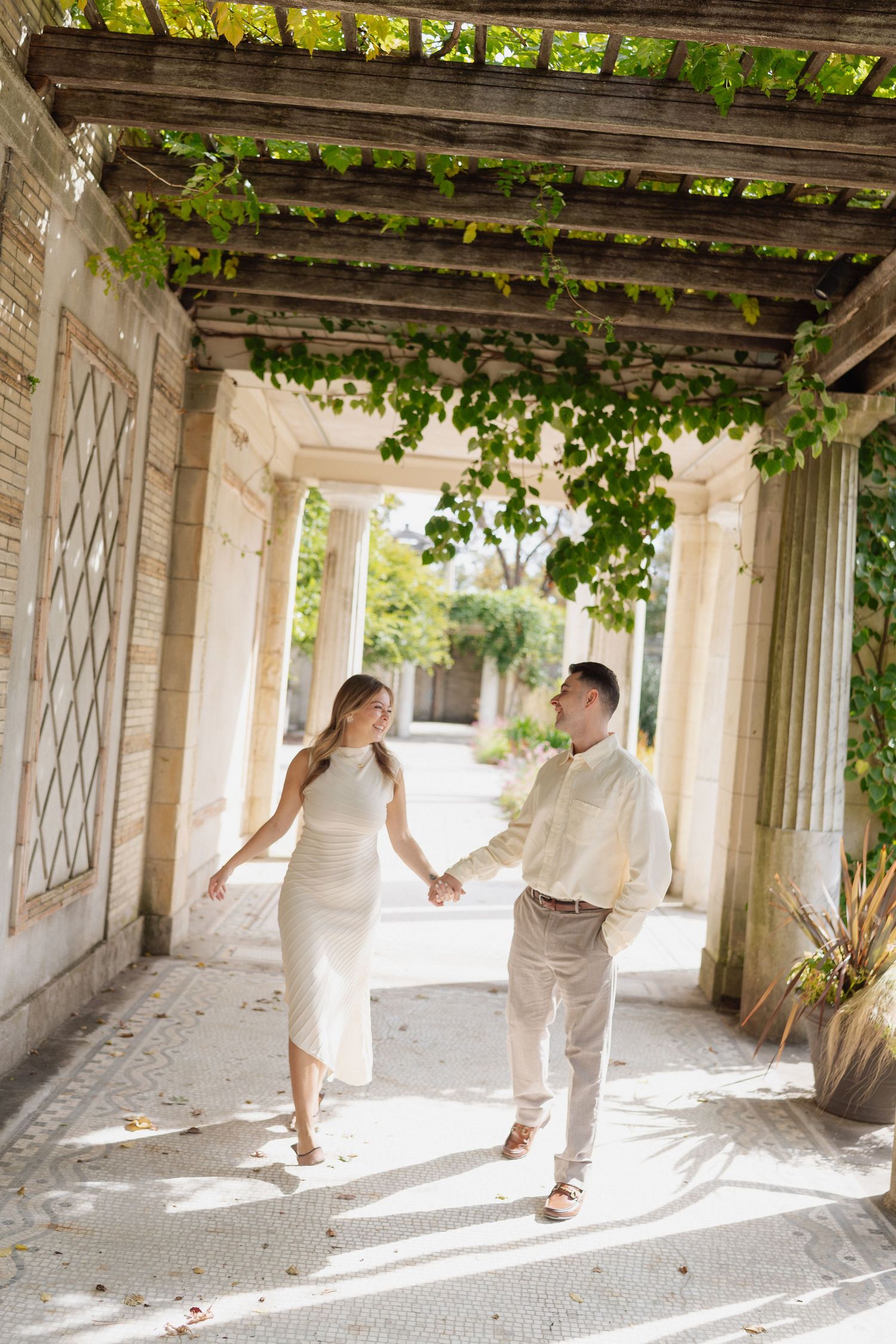 Couple holding hands and walking through a sunlit stone colonnade covered in green vines, woman in white dress, man in linen shirt.
