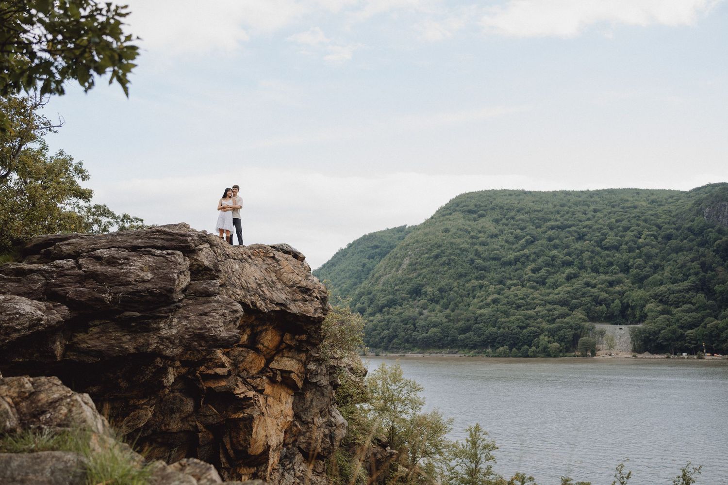 Couple embracing on a rocky cliff edge overlooking a calm river and lush green forested mountains under a soft cloudy sky.