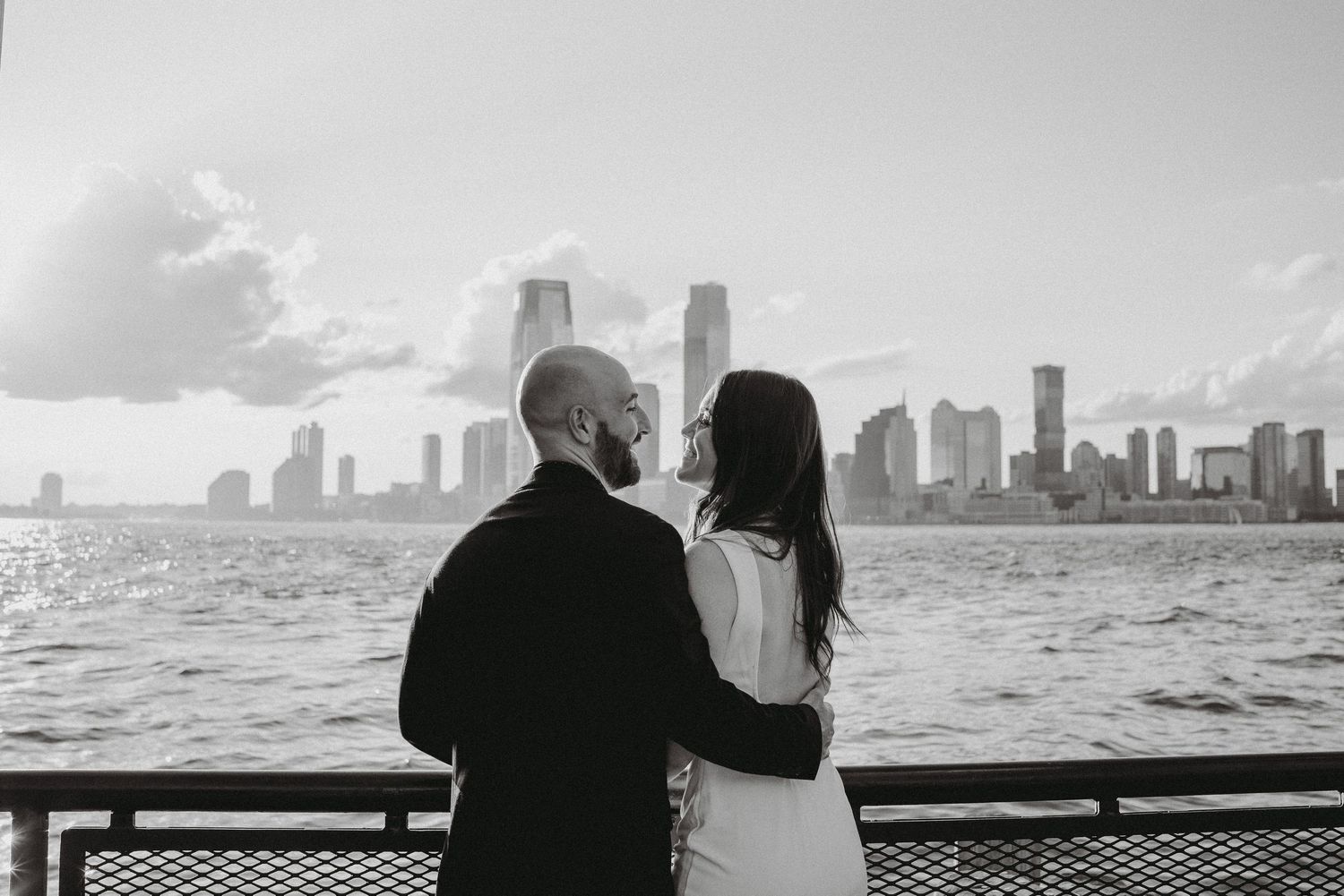 Black and white photo of couple facing the Jersey City skyline across the Hudson River.