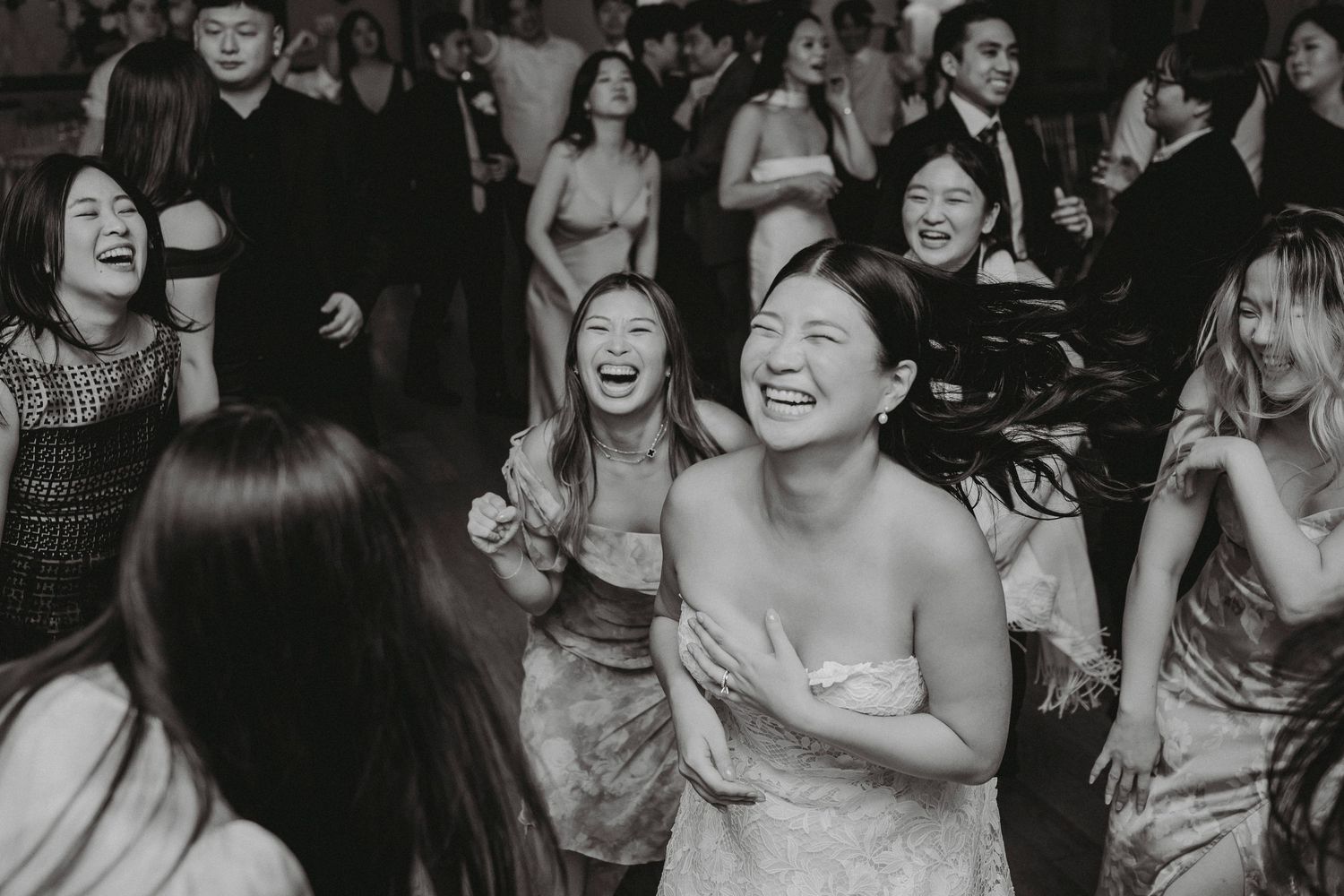 Bride and wedding guests laugh and dance joyfully on the dance floor in black and white reception photo.