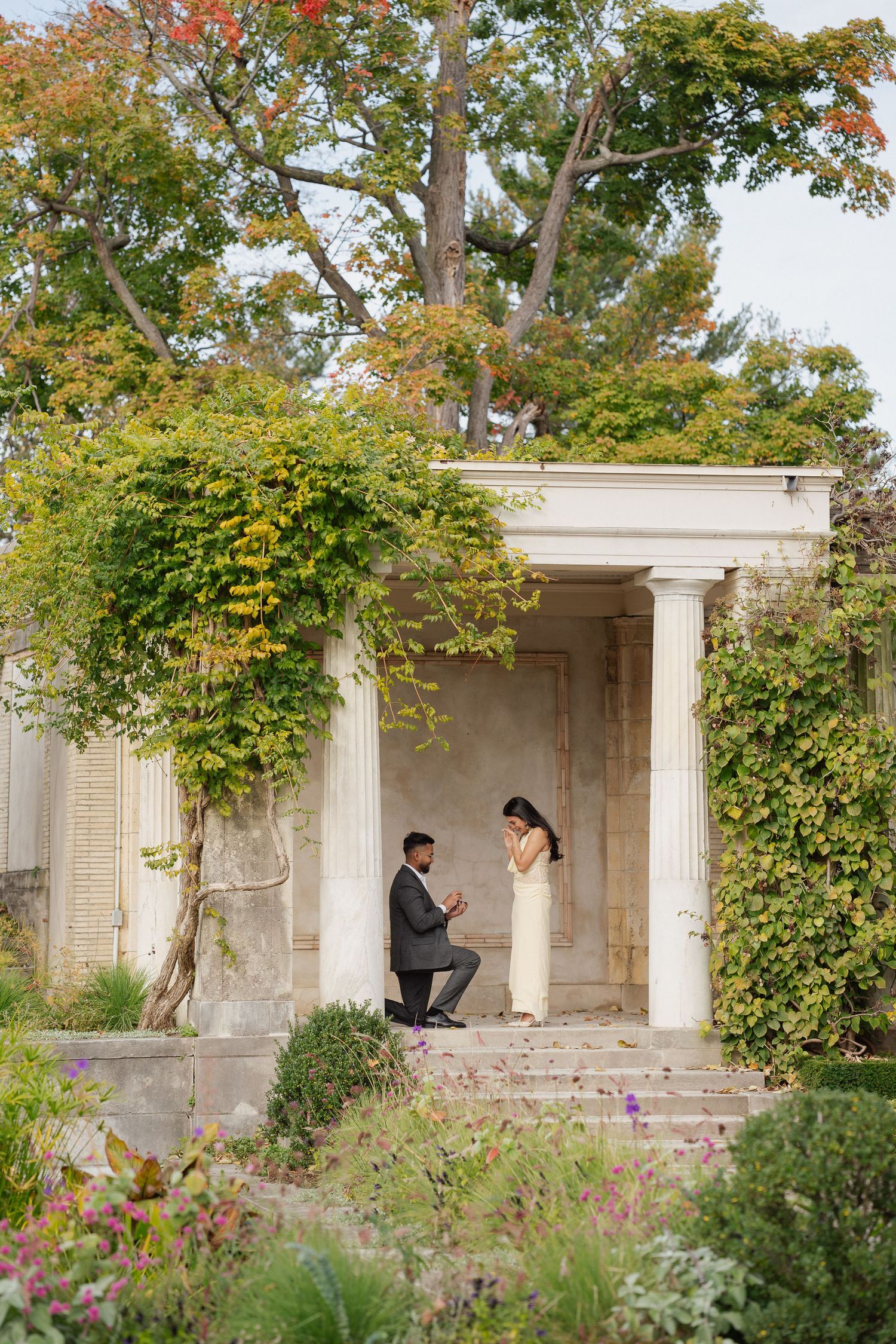 Man in a suit kneels on one knee proposing to a woman in a long white dress beside a classical stone pavilion surrounded by autumn foliage.