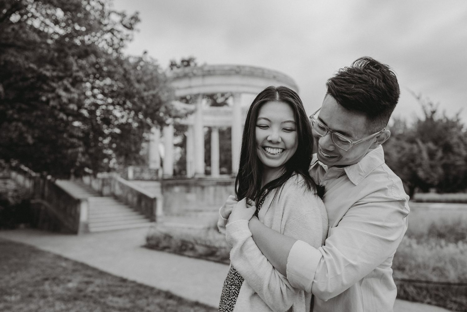 Black and white engagement photo of a smiling couple embracing outdoors with a classical columned pavilion and trees in the background.