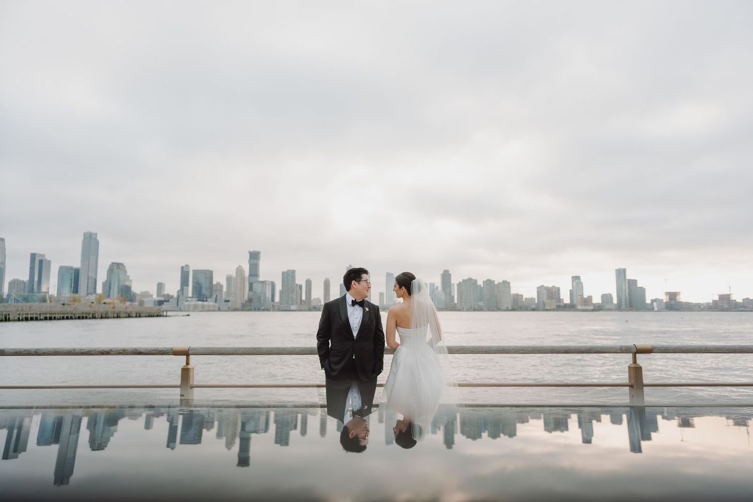 Bride and groom pose by reflective pool with NYC skyline backdrop on their wedding day.