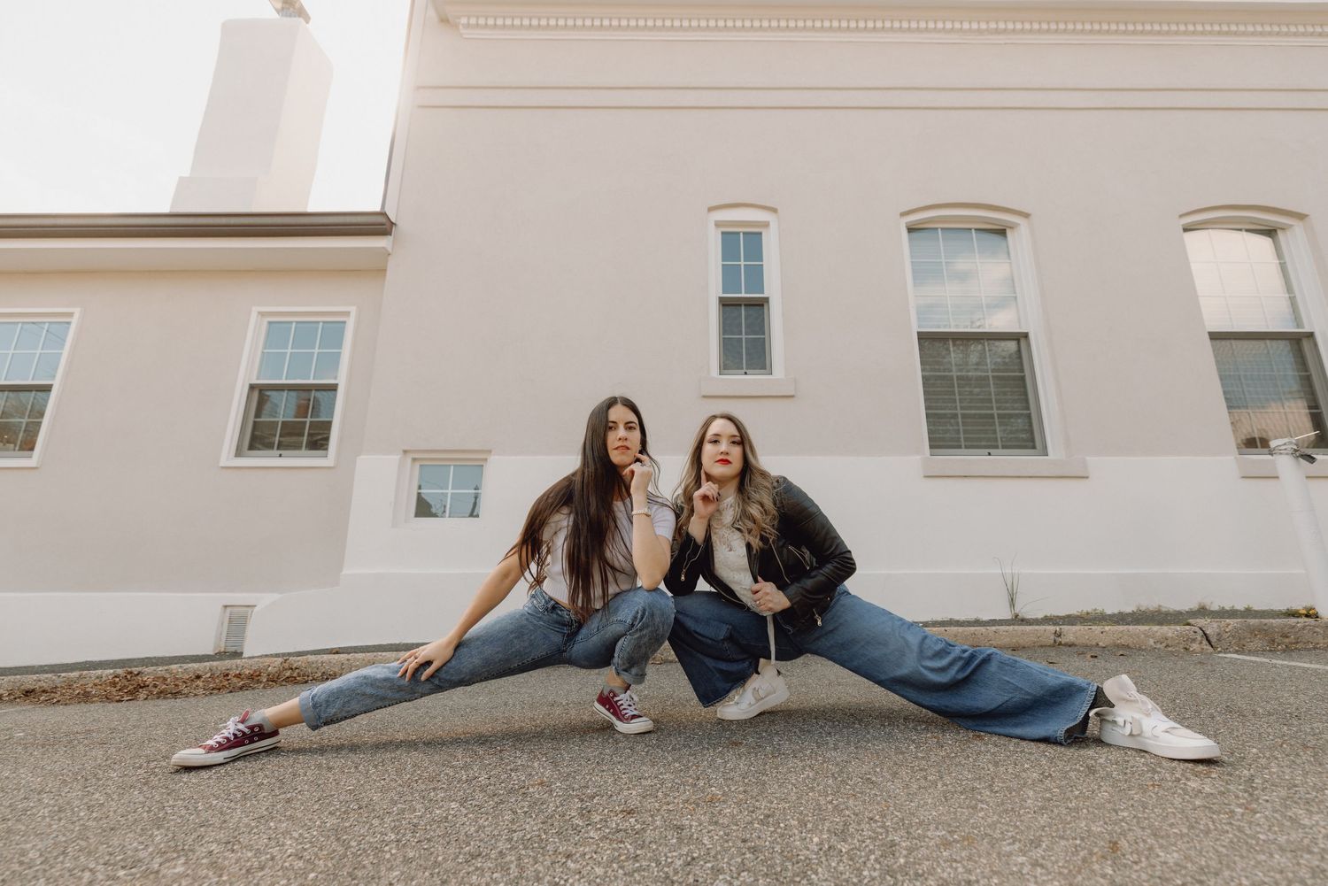 Two young women sitting in a split pose on pavement in front of a white building, wearing jeans and sneakers.
