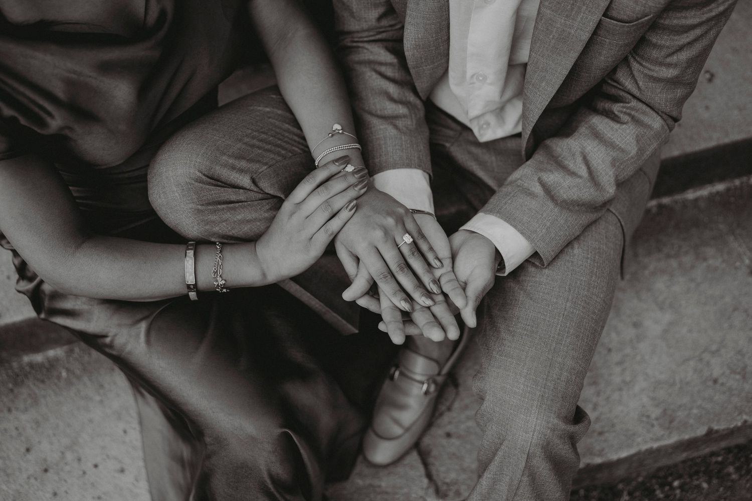 Black and white close-up of couple holding hands showing engagement ring on stone steps.