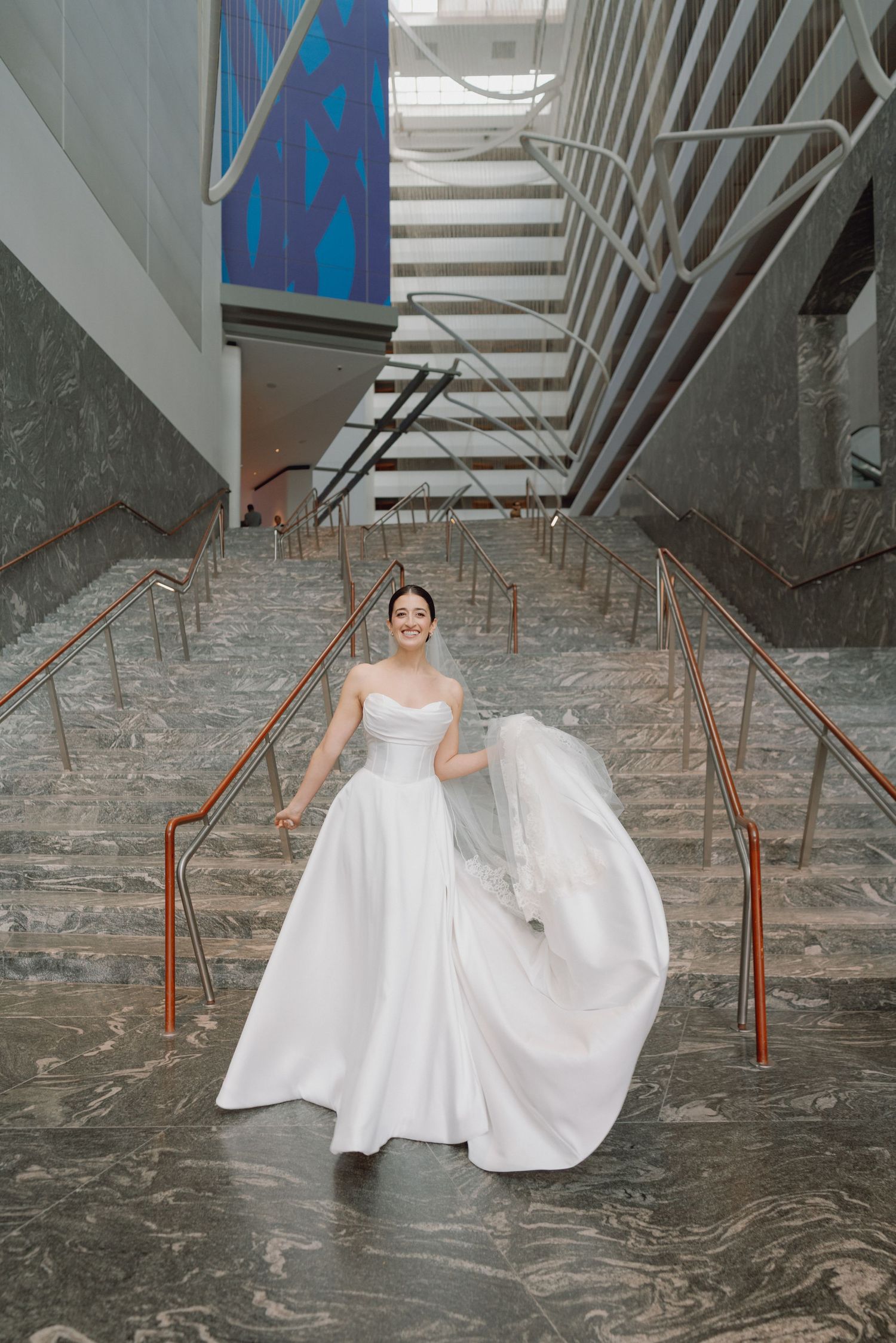 Bride in strapless white gown with flowing veil descending marble stairs in modern venue.