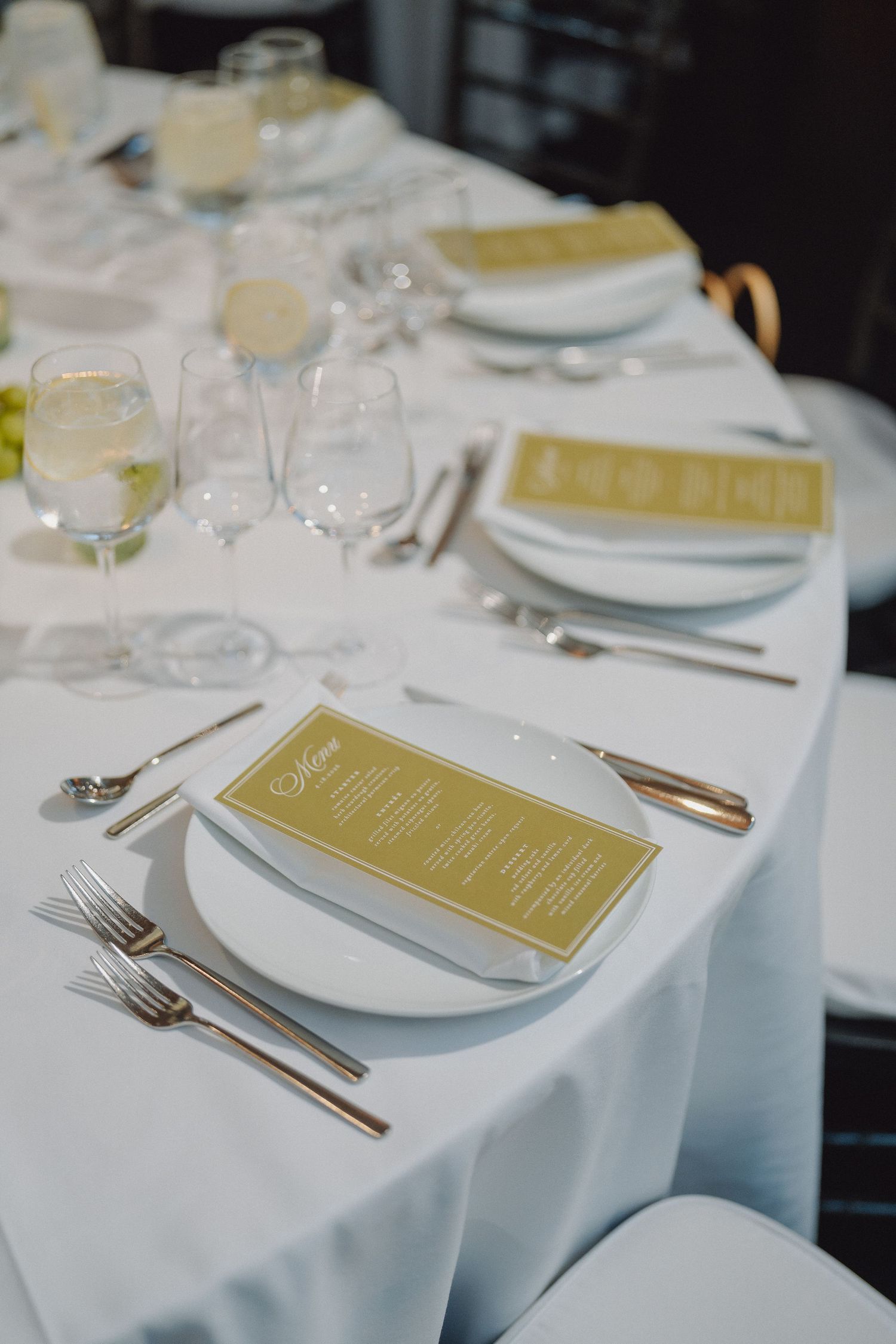 Elegant wedding reception table setting with white linens, gold menus, crystal glasses, and silver cutlery.