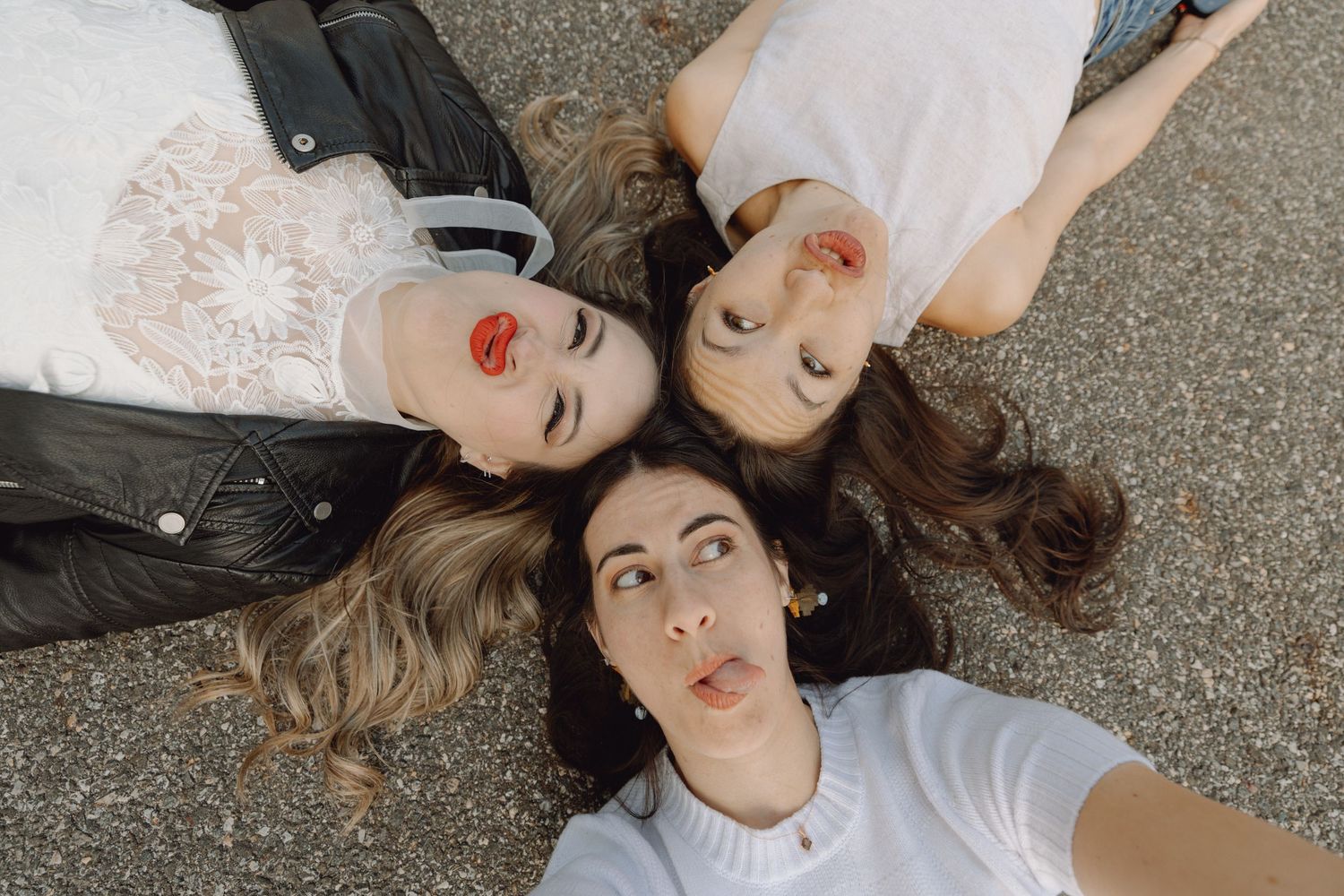 Four women lying in a circle on pavement with heads together taking a selfie from above.