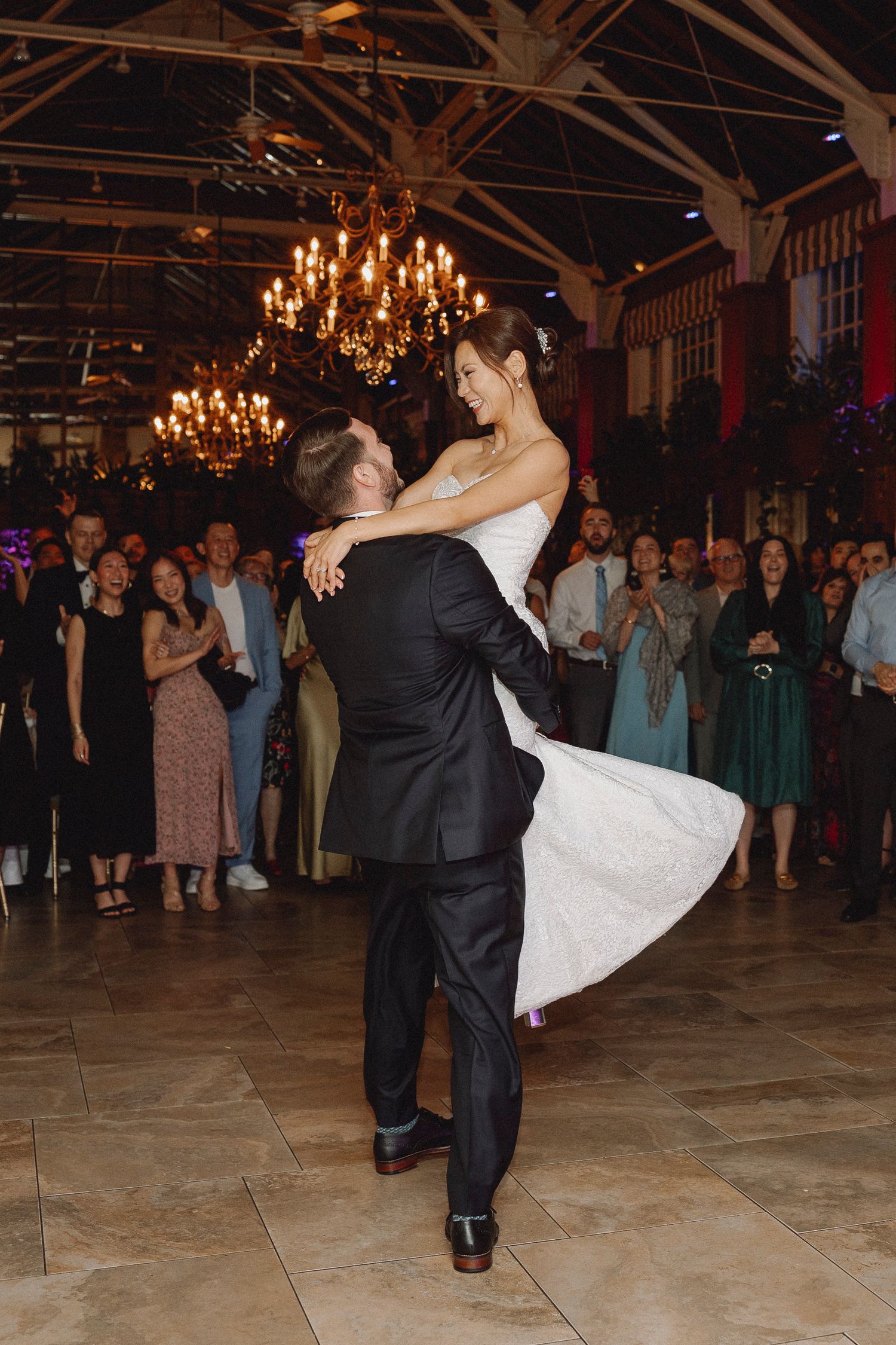 Groom lifting bride during first dance surrounded by cheering wedding guests.