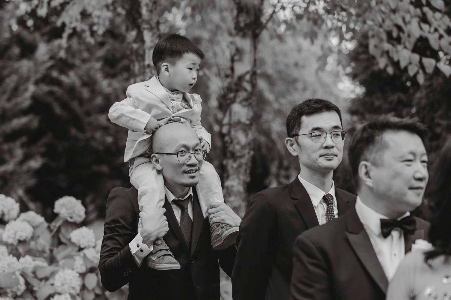 Man carries young child on shoulders alongside wedding guests in black and white outdoor ceremony photo.