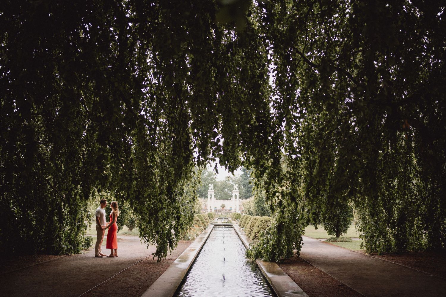 A couple stands together along a reflecting pool beneath a dramatic canopy of weeping trees.