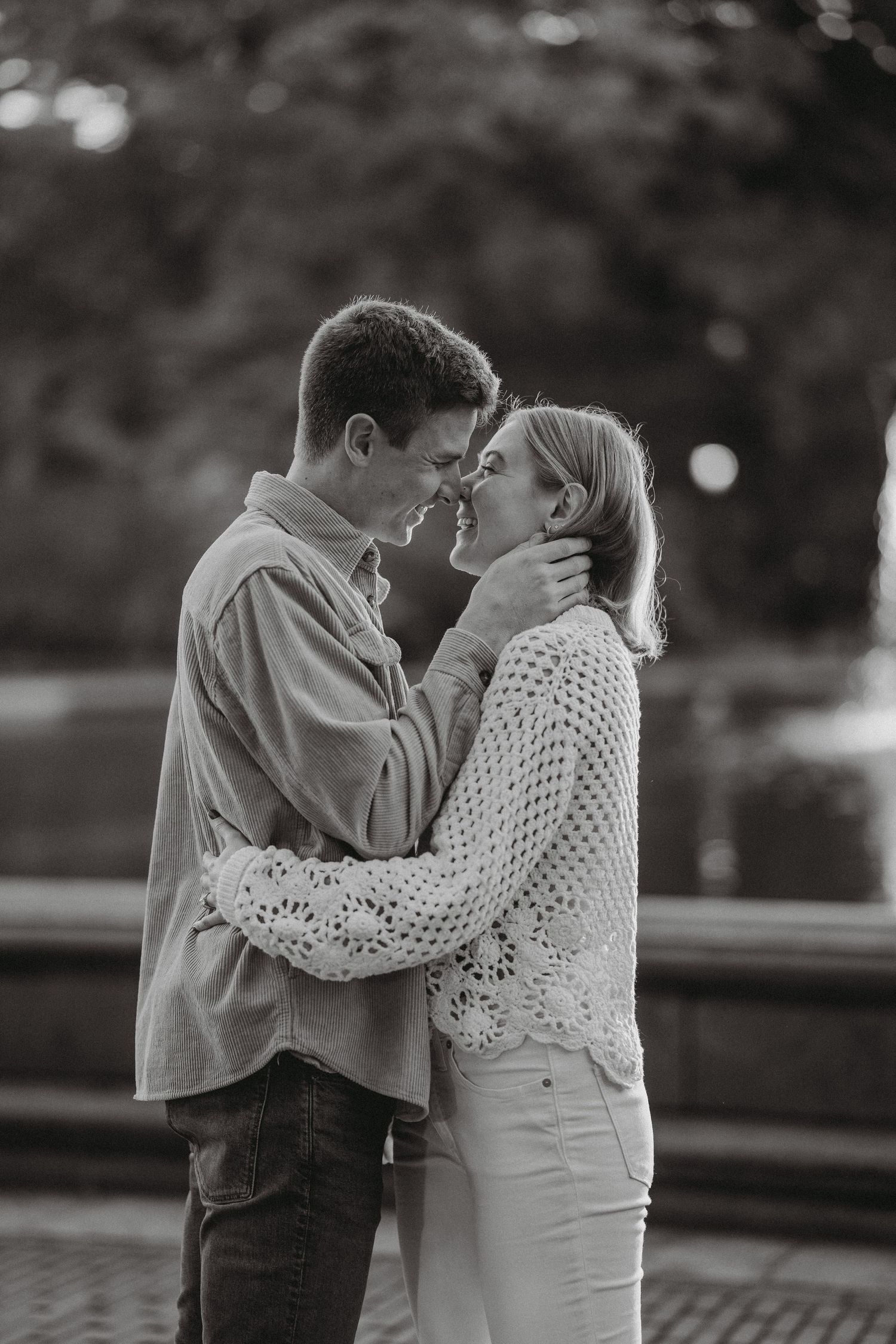 Black and white photo of a young couple almost kissing while embracing outdoors in a park setting.
