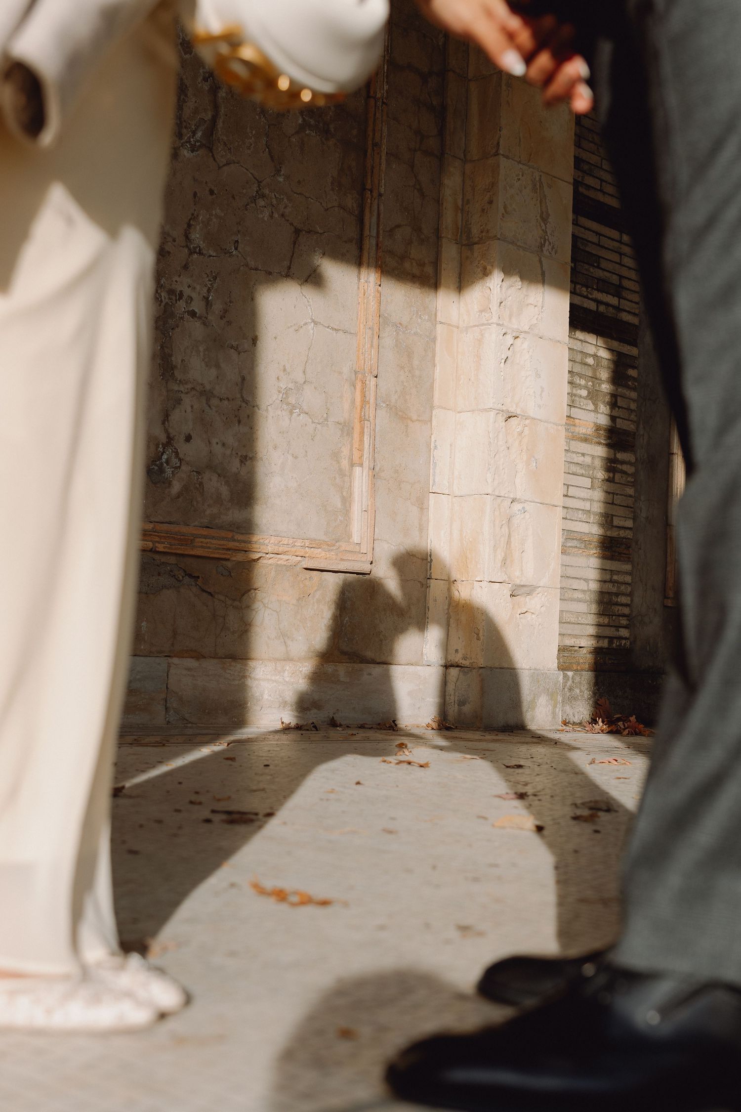 Two people stand facing each other on a sunlit stone pavement, their shadows cast on an old weathered wall behind them.