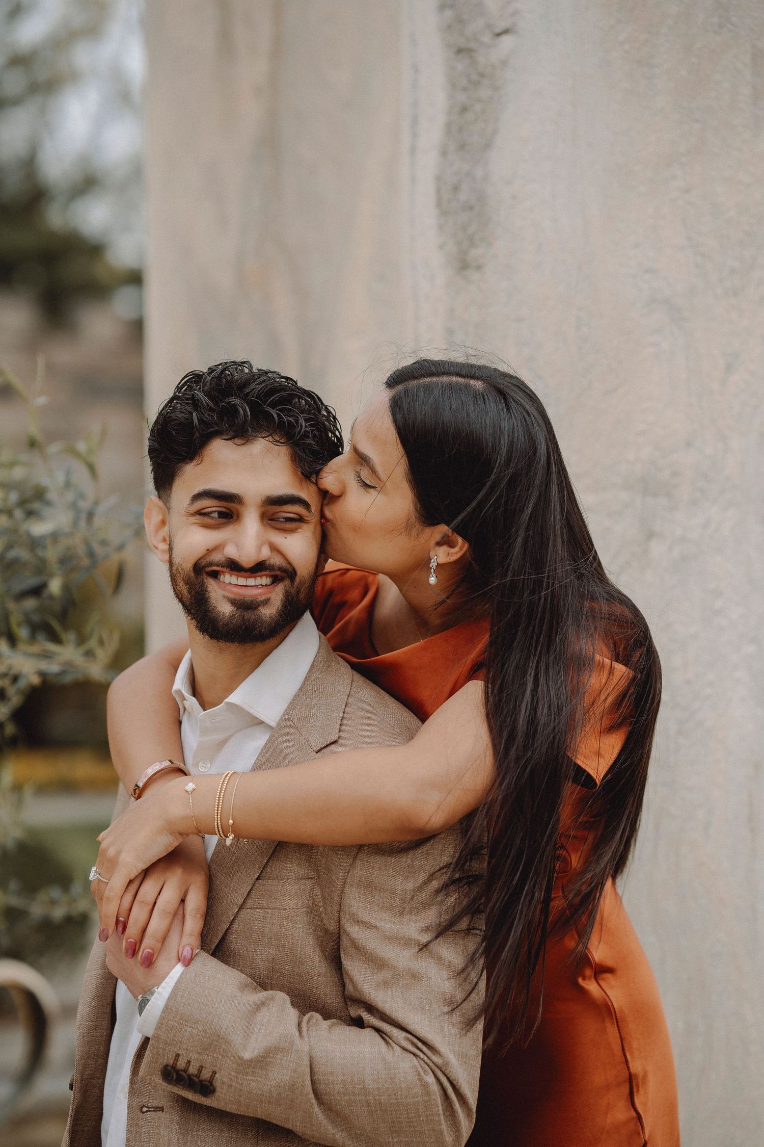 Couple embracing near stone wall, woman kissing mans cheek, man smiling, wearing beige suit and rust orange dress.