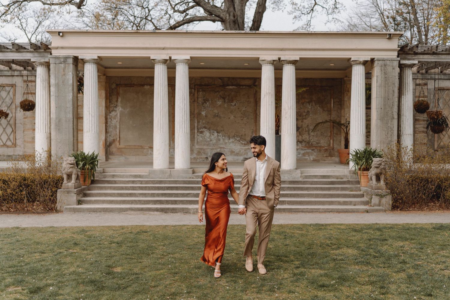 Couple walking hand in hand on grass in front of a classical columned stone pavilion.