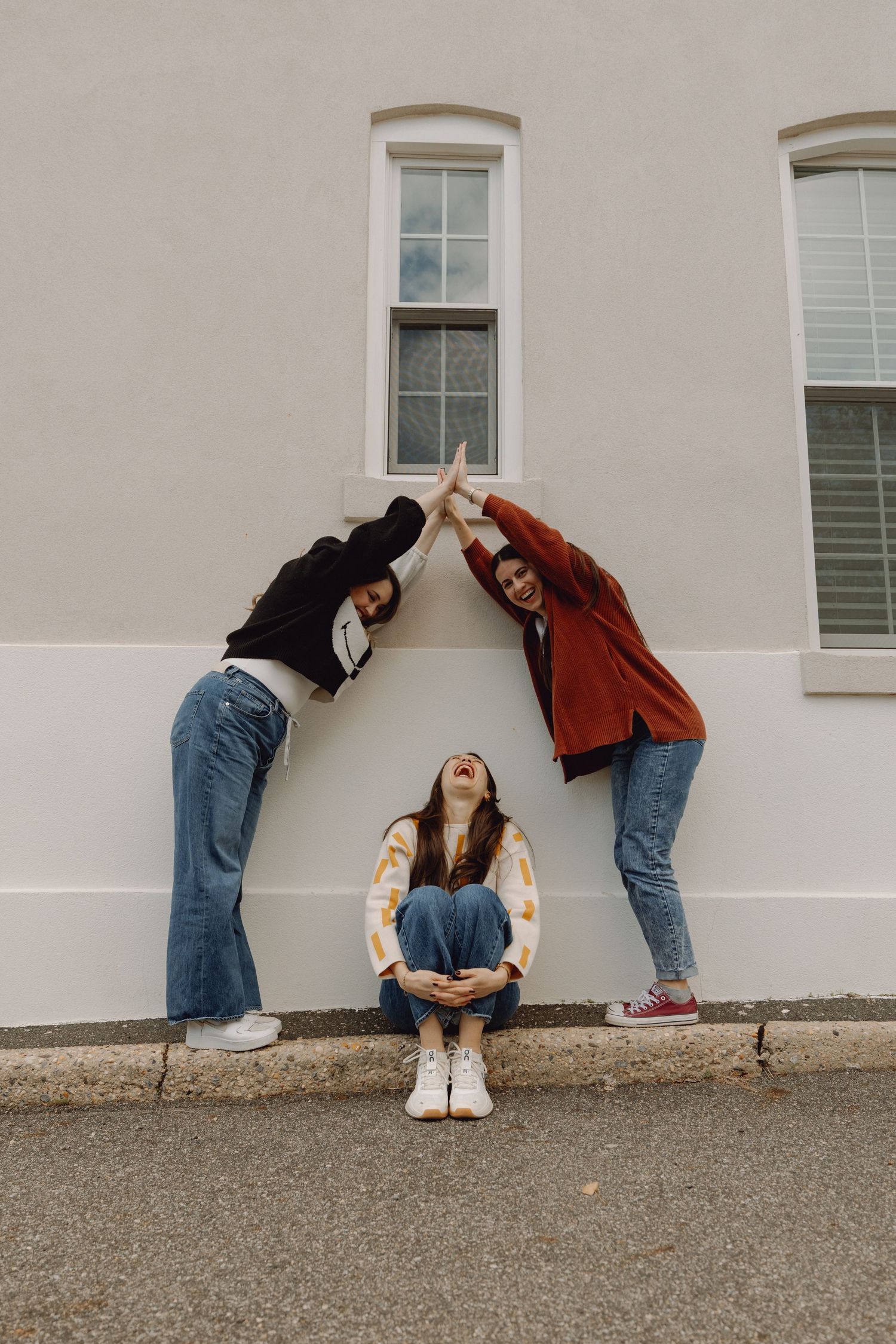 Three friends pose playfully against a building, two forming a roof shape while one sits laughing beneath them.