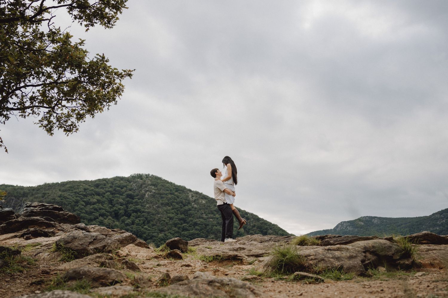 Couple embracing and lifting each other on a rocky overlook with green forested hills and a cloudy sky behind them.