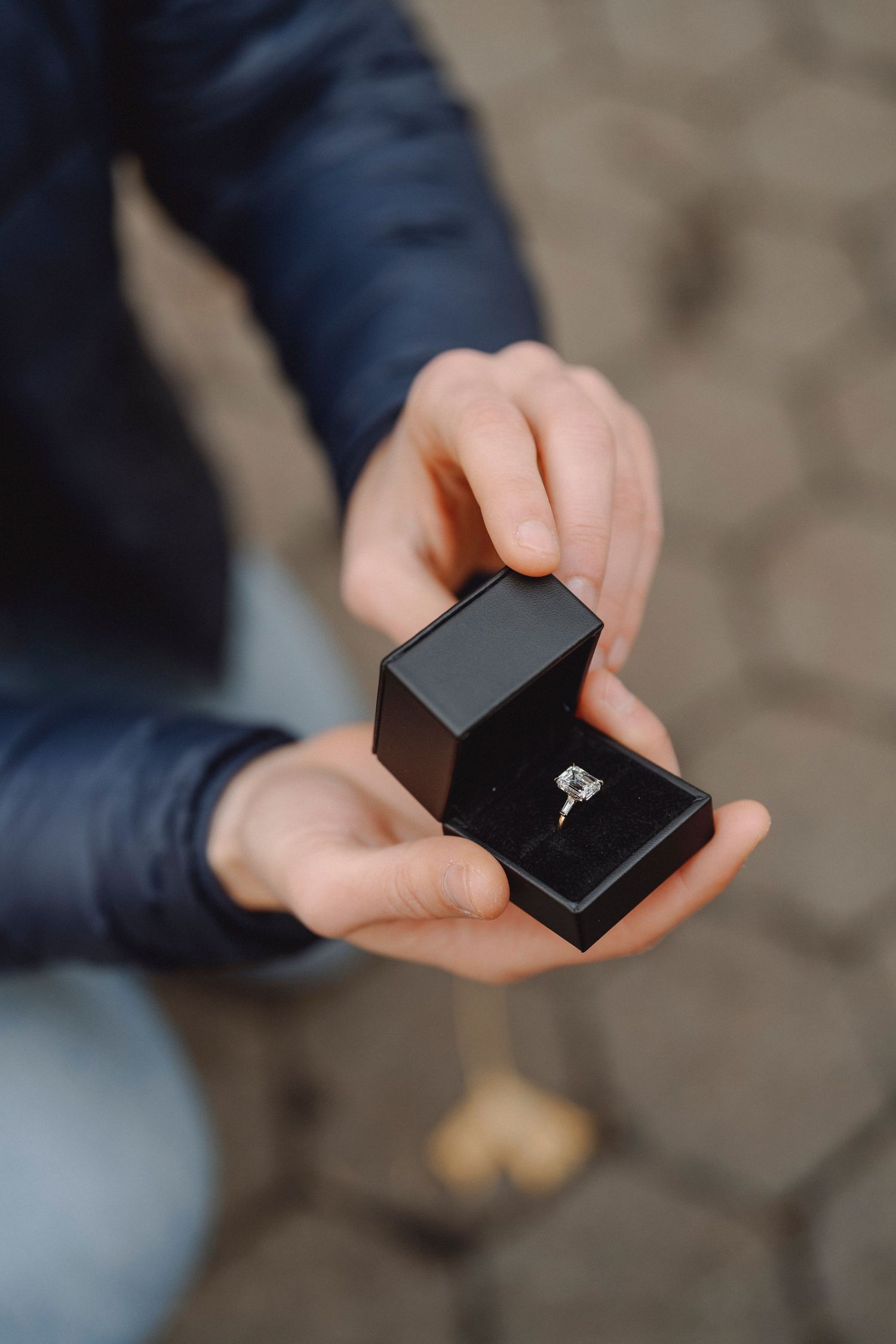 Person kneeling in a navy jacket holds an open black box with a sparkling diamond engagement ring, ready to propose on a cobblestone street.